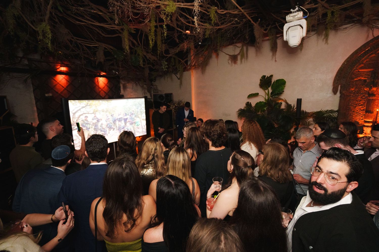 Large group of event attendees watching a presentation in an indoor venue with hanging plants.