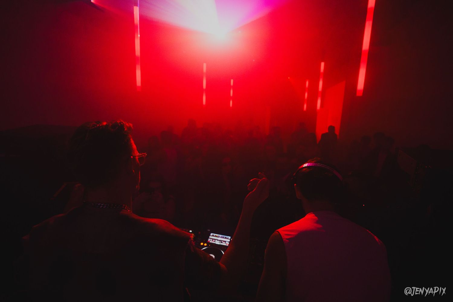 DJs performing at a DJ booth under dramatic red lighting at a nightclub event.