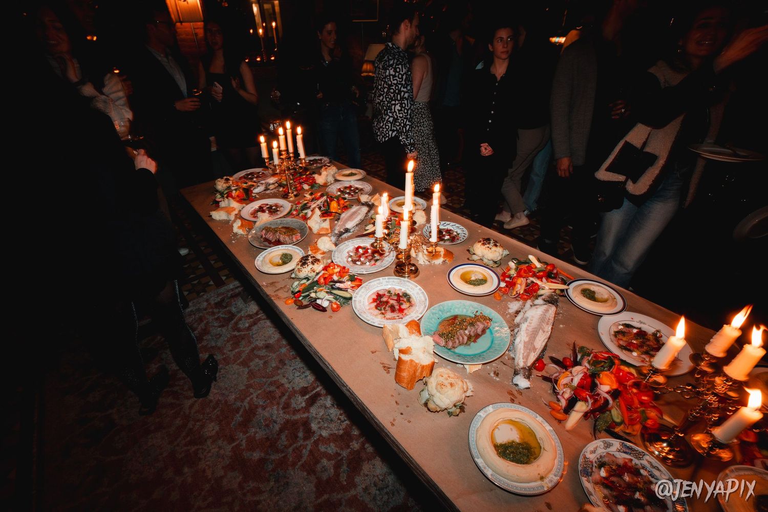 Candlelit table set with multiple dishes and plates in a dark atmospheric setting.