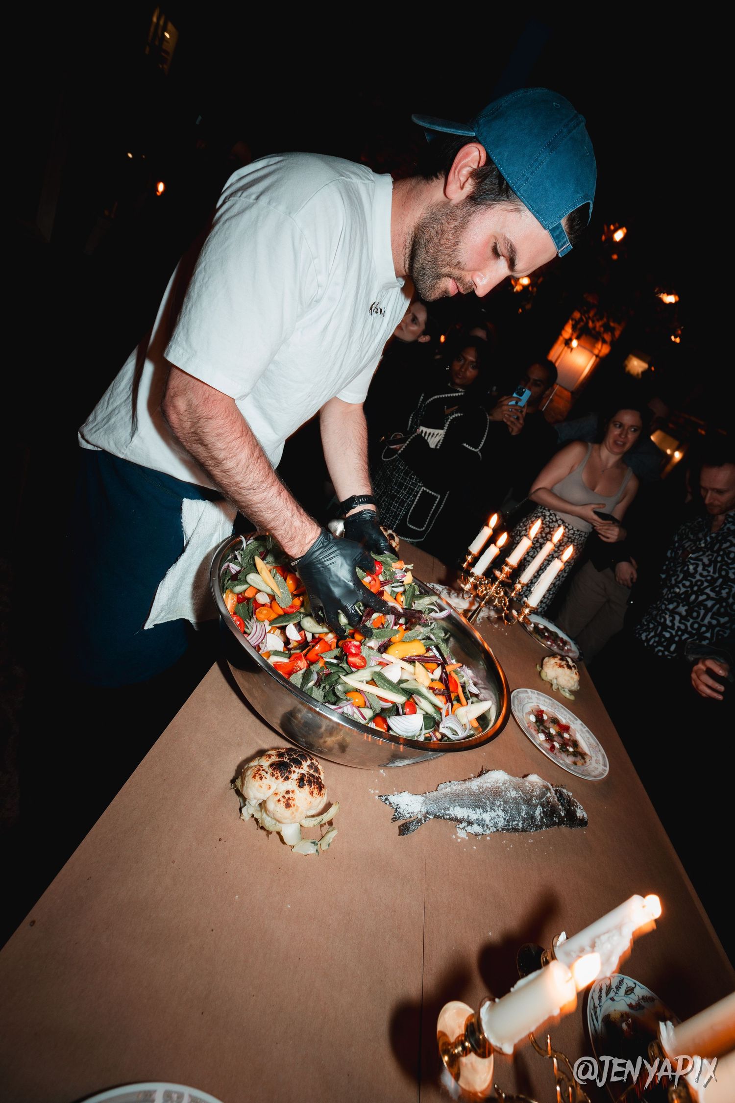 A chef in white plating food on a table with candles and decorative dishes.