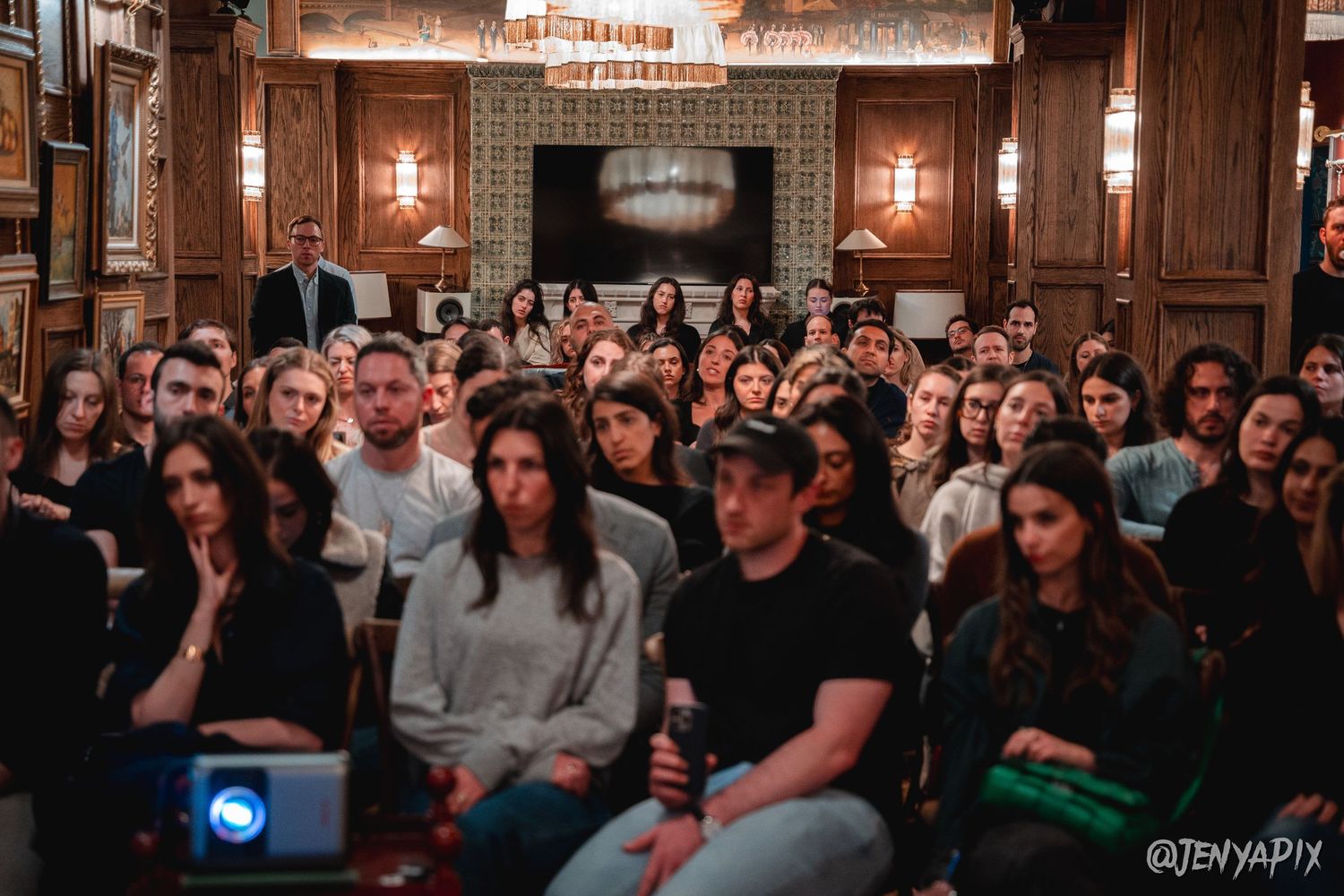 Full audience in wood-paneled venue with vintage lighting and fireplace.