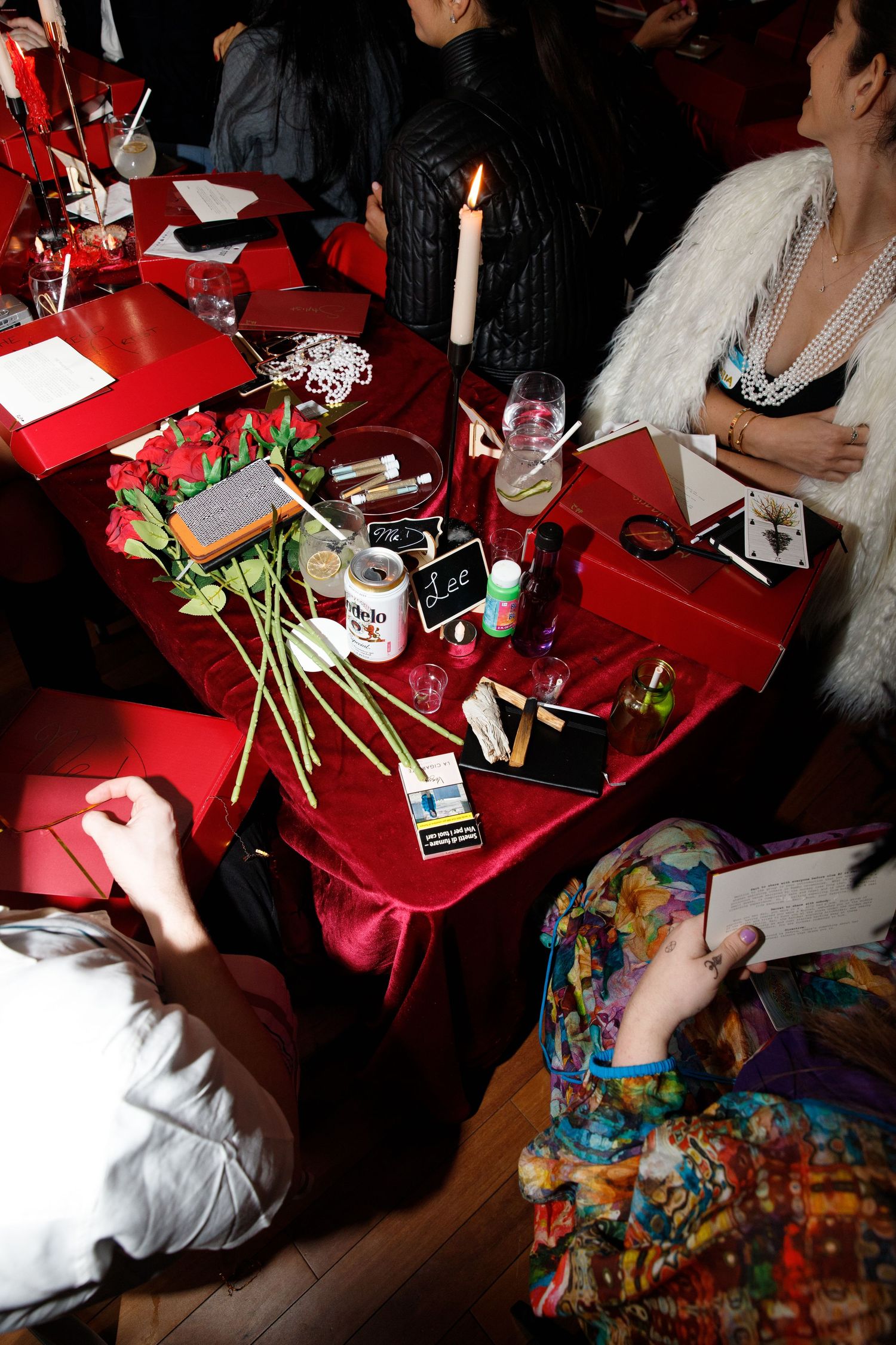 A dark room with a red tablecloth covered table featuring drinks, flowers and party supplies.