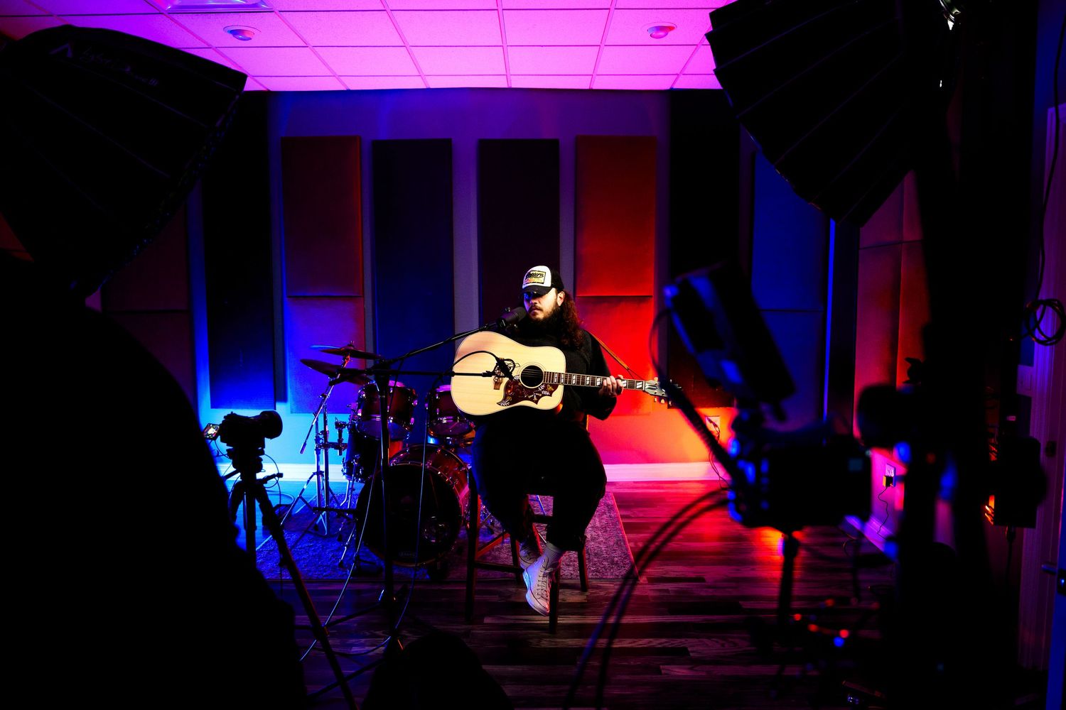 Musician performing with acoustic guitar on stage lit by red and blue lights.