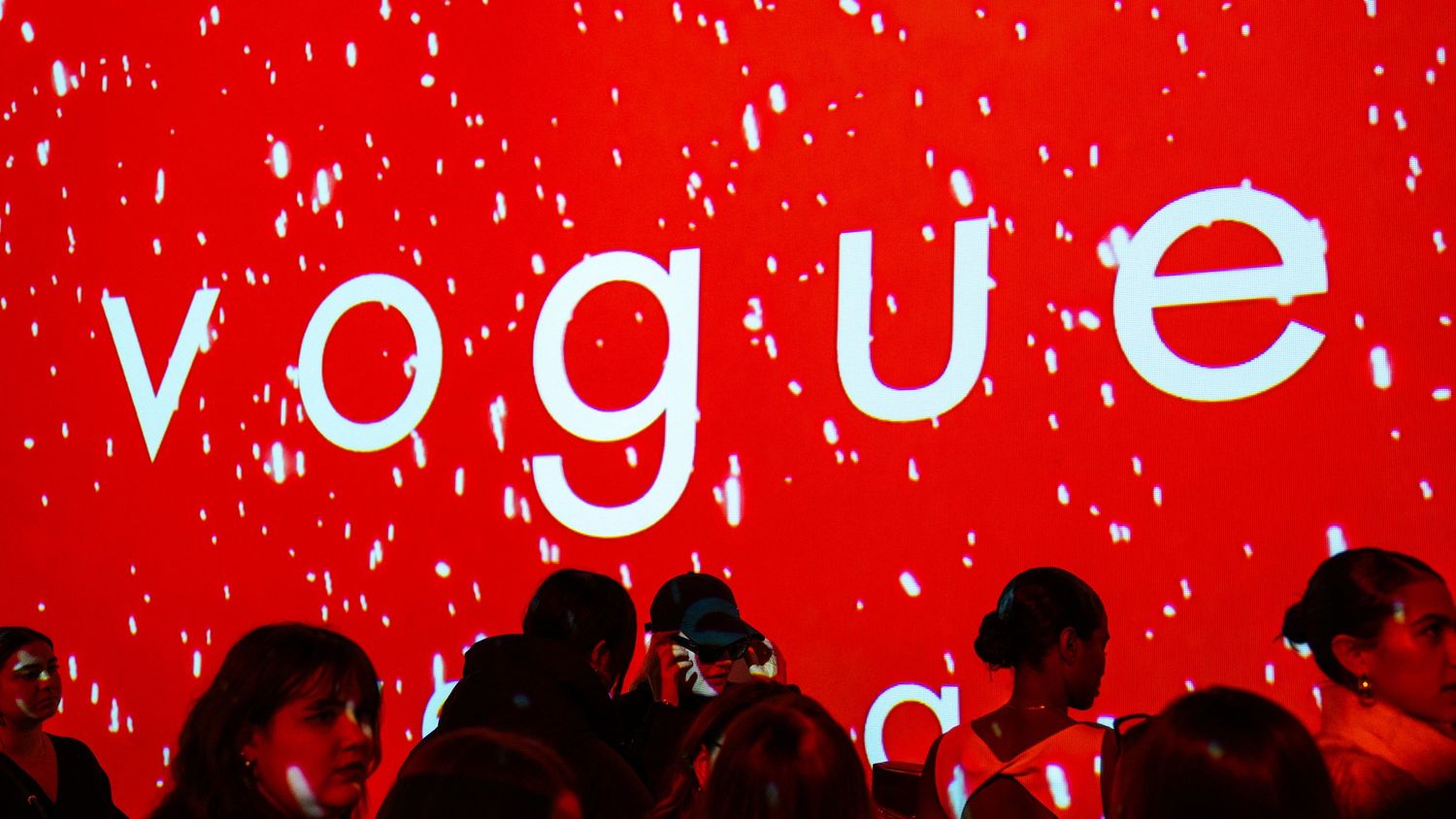 Vogue logo projected on red wall with snow effect and audience silhouettes.