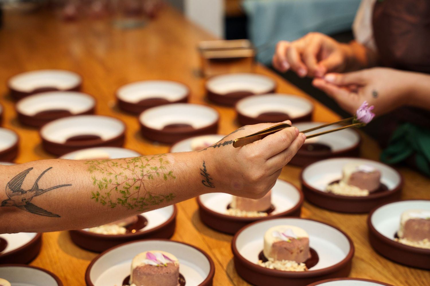 Tattooed hands using chopsticks to garnish plated desserts on a wooden restaurant table.