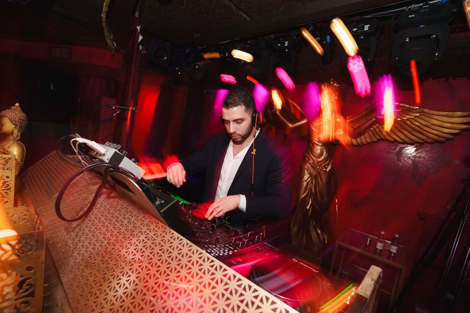 DJ performing behind decorative booth with colorful lighting effects in nightclub atmosphere.