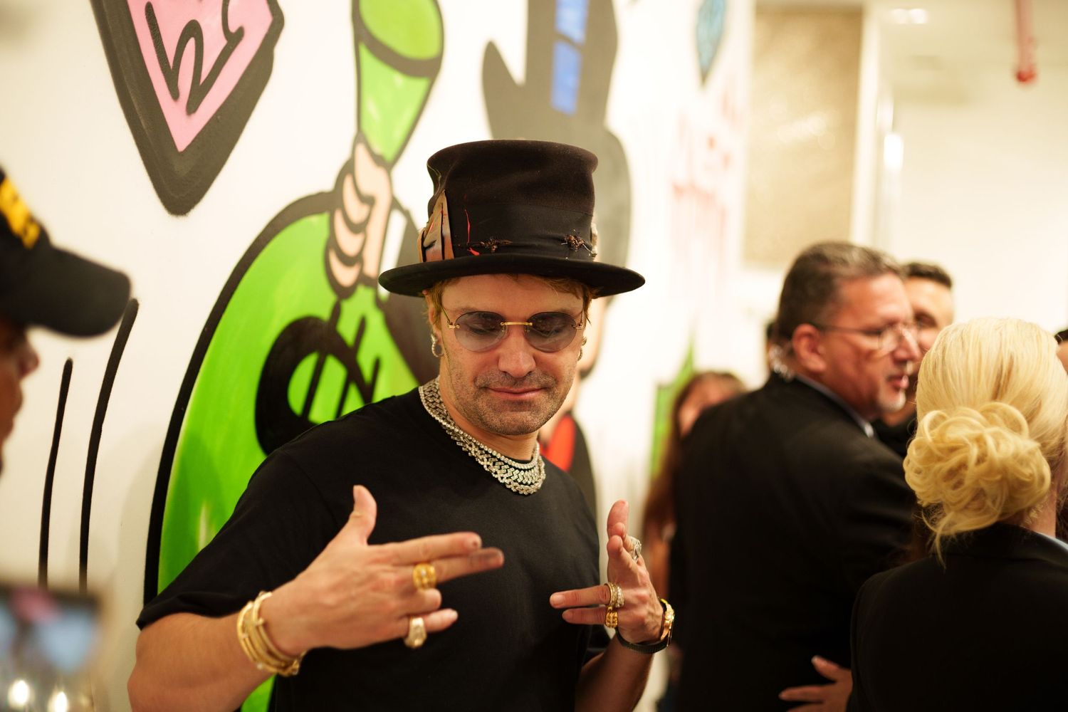 Person in top hat and glasses gesturing at art gallery event.