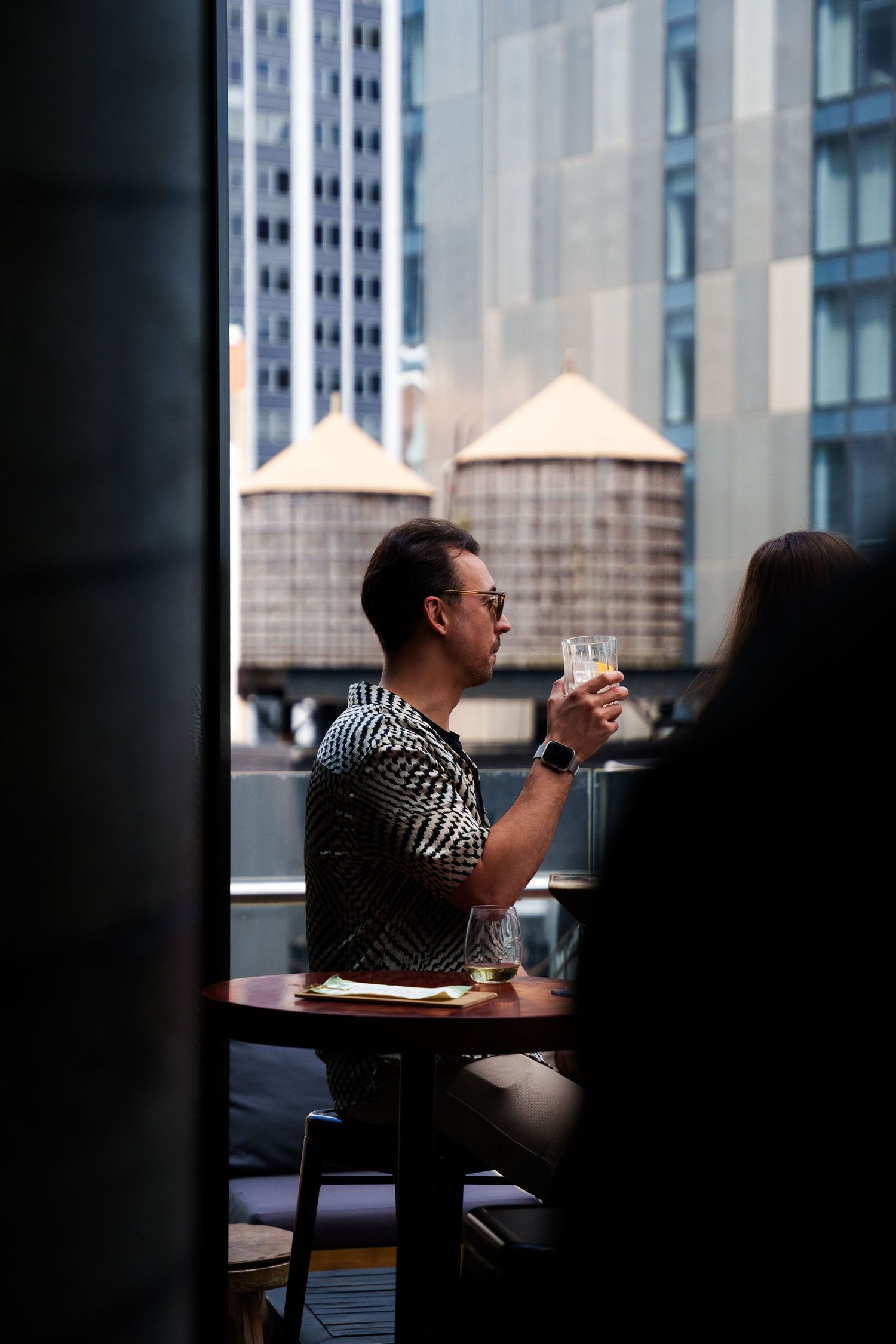 Person enjoying a drink at outdoor cafe with city buildings in background.