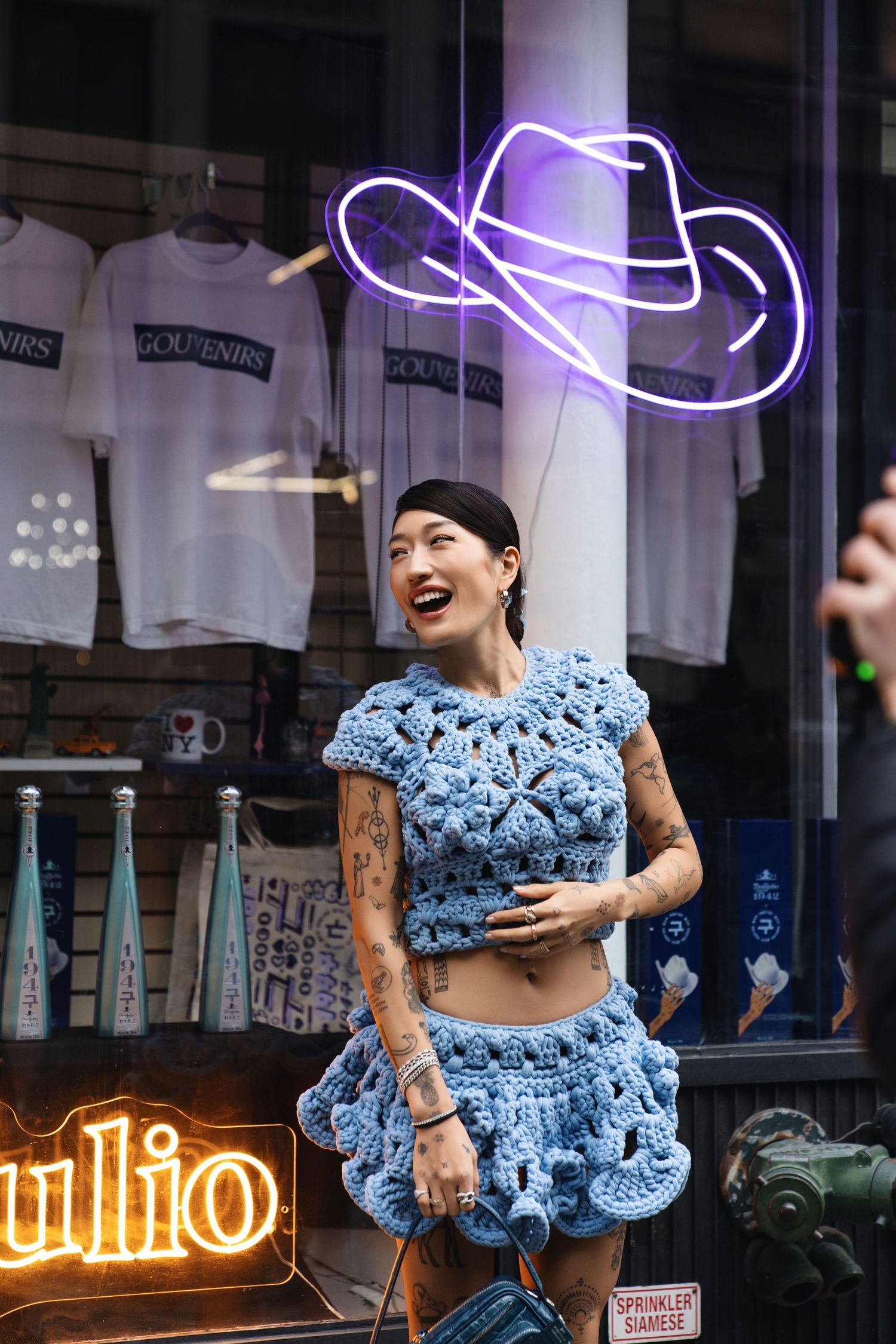 Person in blue crochet outfit laughing under purple neon cowboy hat sign.