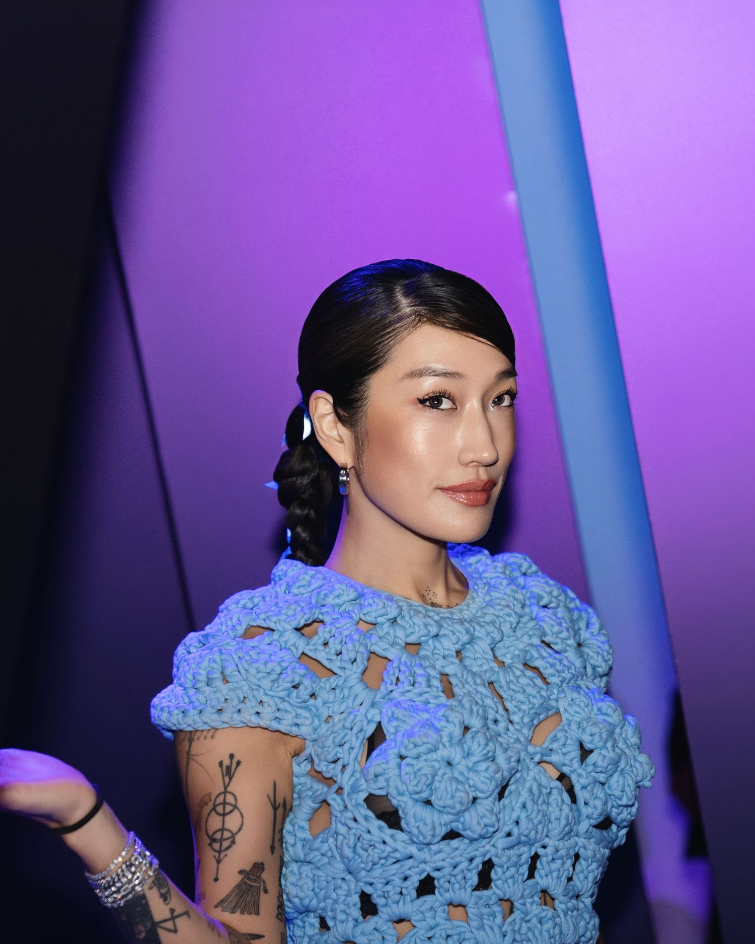 Portrait shot of person in blue crochet top with visible tattoos against purple lighting.