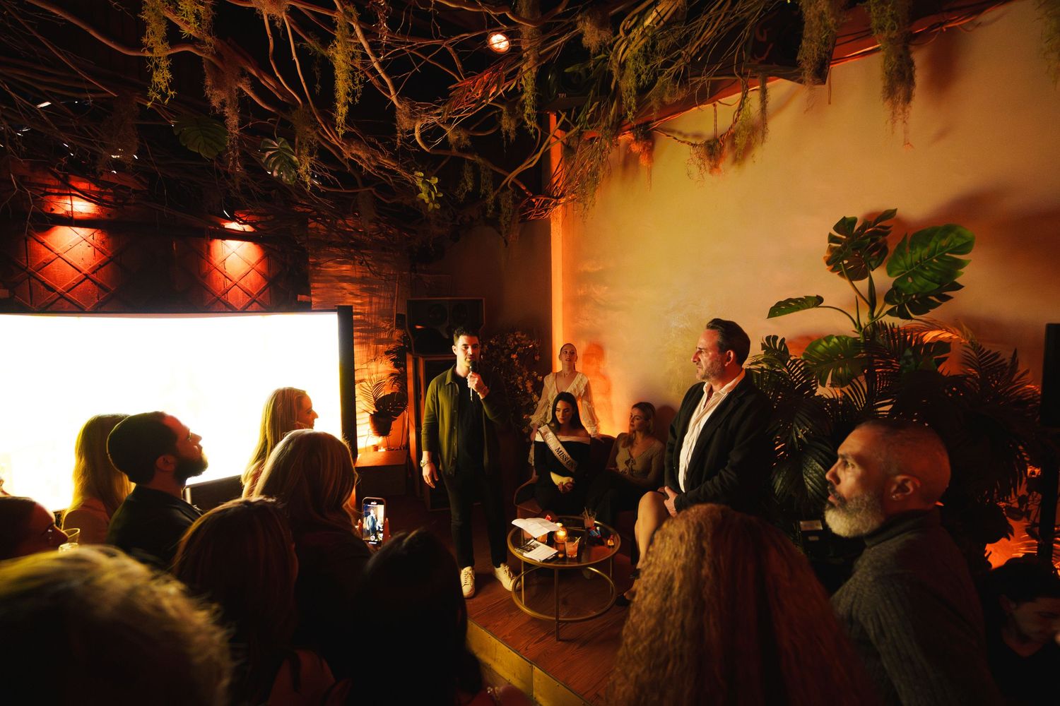 Crowd gathered in a warmly lit venue with greenery and decorative lighting for an evening performance.