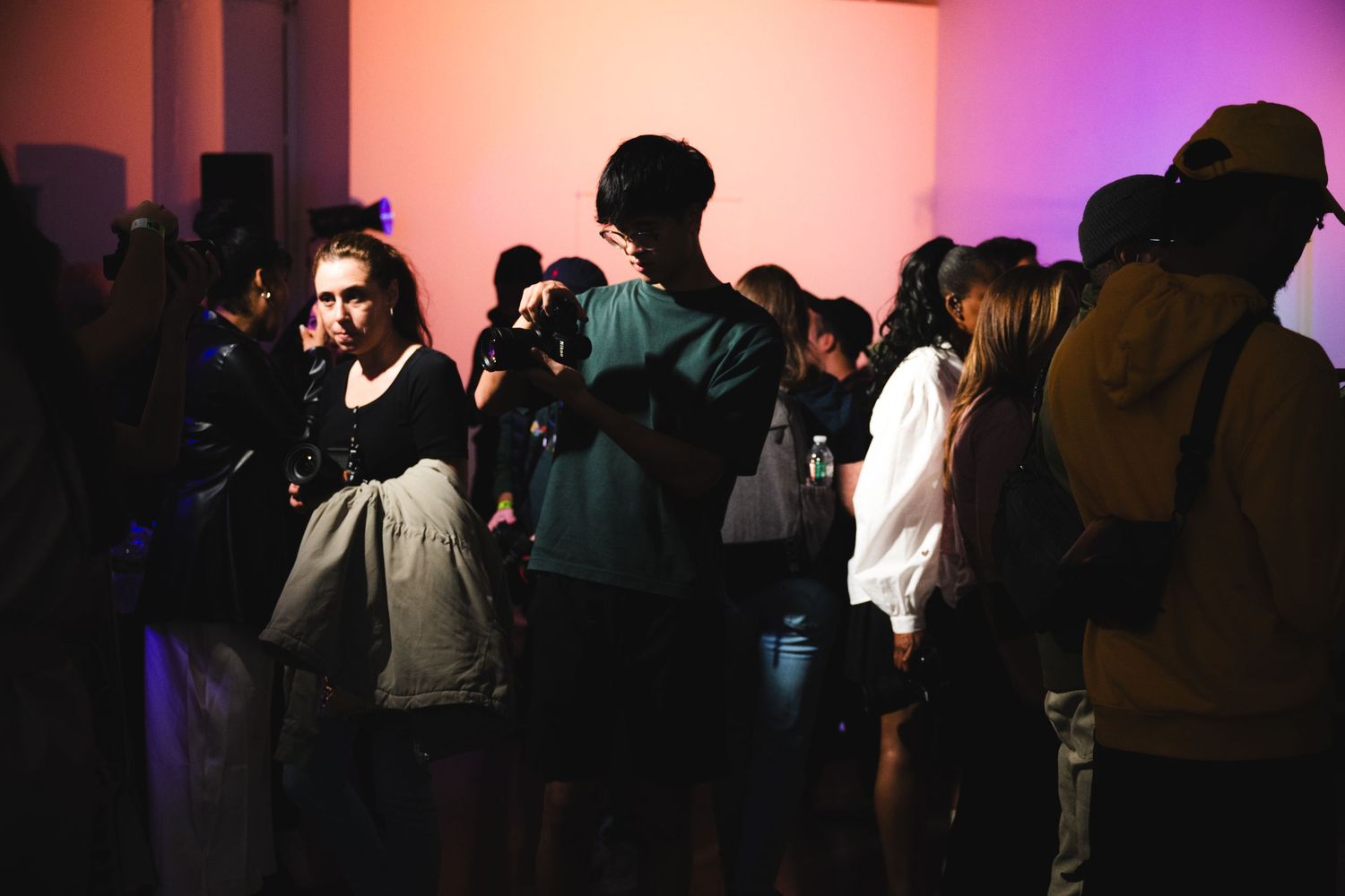 A dimly lit crowd of people attending an indoor event with pink and purple lighting.