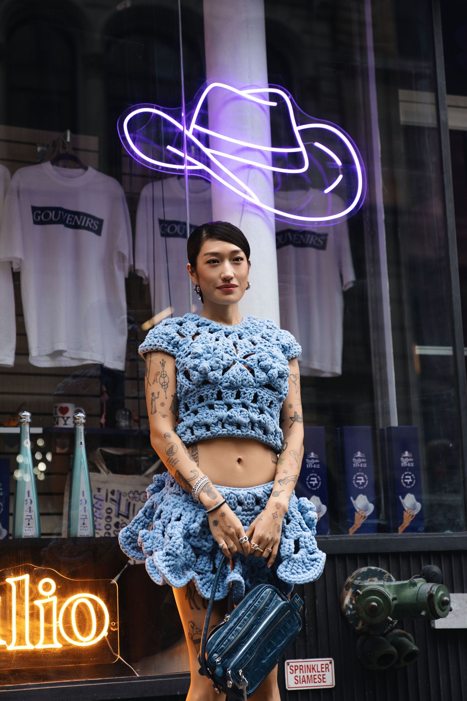 Blue crochet crop top and skirt ensemble photographed under neon cowboy hat sign at storefront.