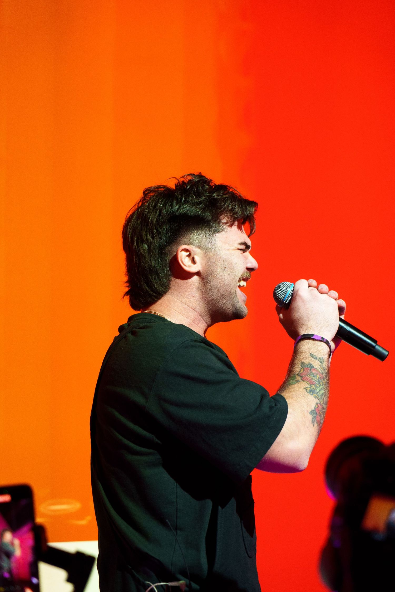 Performer singing passionately into microphone against vibrant orange-red backdrop during live show.