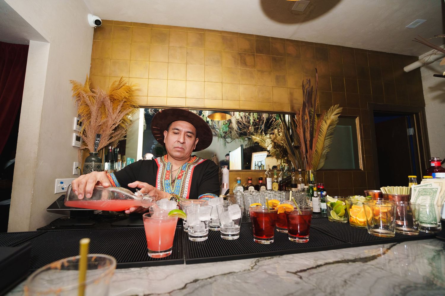 Bartender in a hat pouring colorful cocktails behind a well-stocked bar.