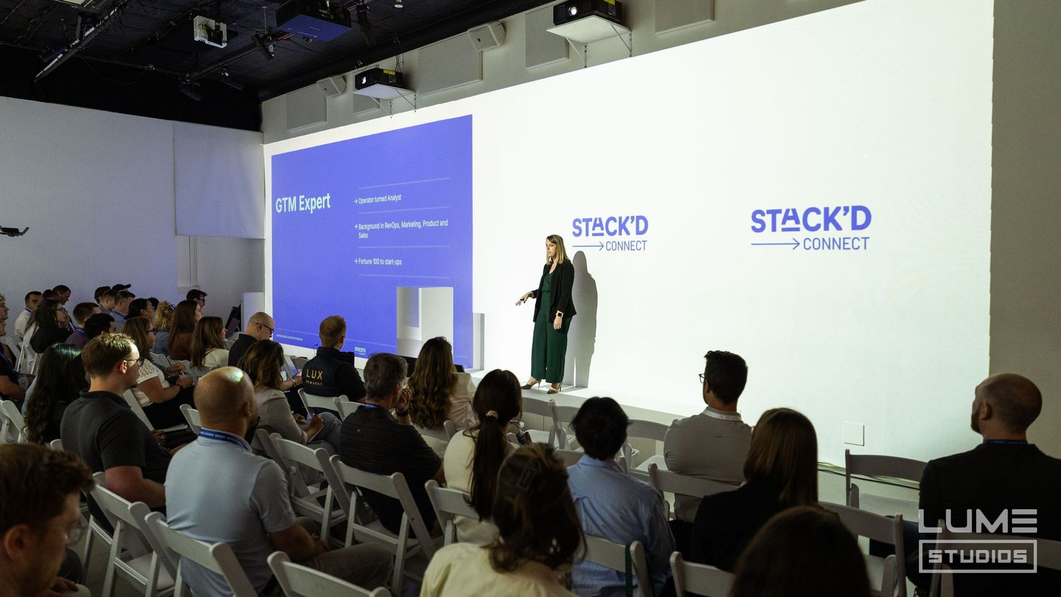 Business presentation with audience seated in rows watching speaker present on large white screen with company logos.