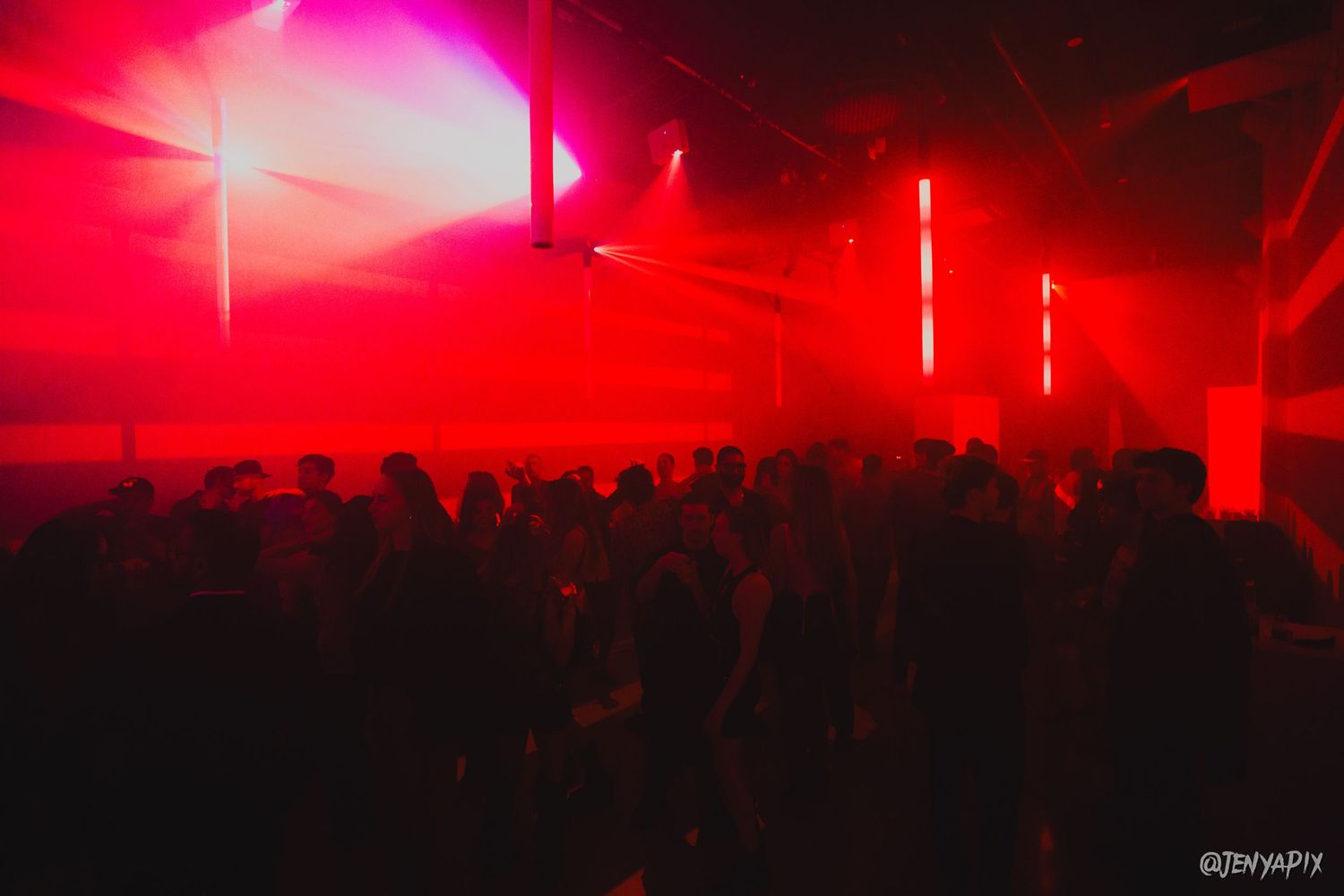 A crowded nightclub dance floor illuminated by intense red lighting and vertical light beams.