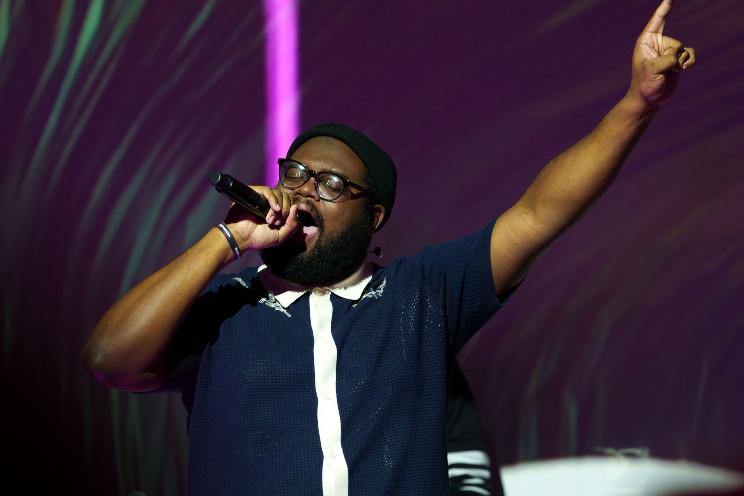 Energetic performer in navy outfit raises arm while singing into microphone against purple backdrop.