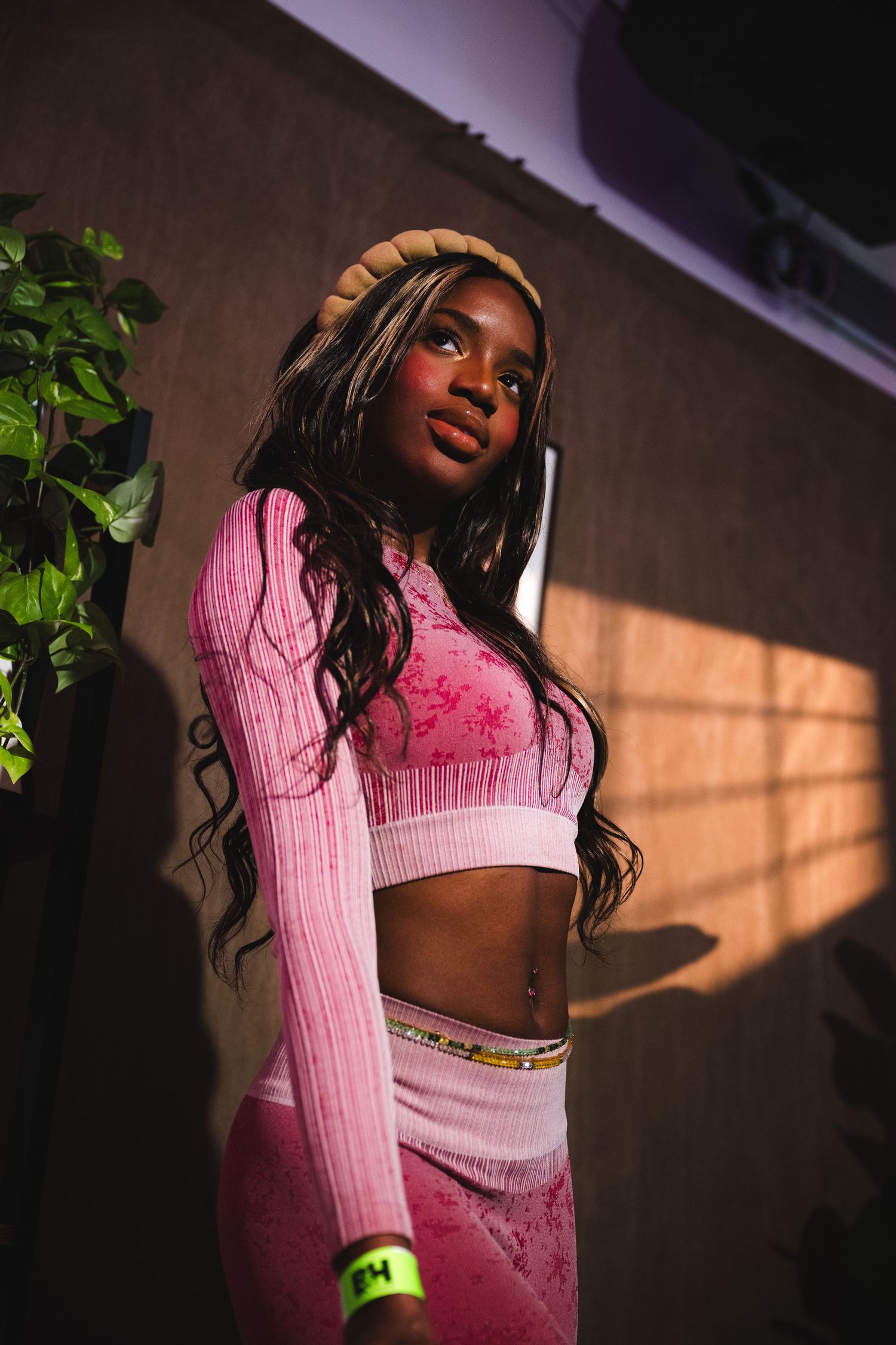 A model in pink knit outfit poses against a wall with dramatic lighting and plants.
