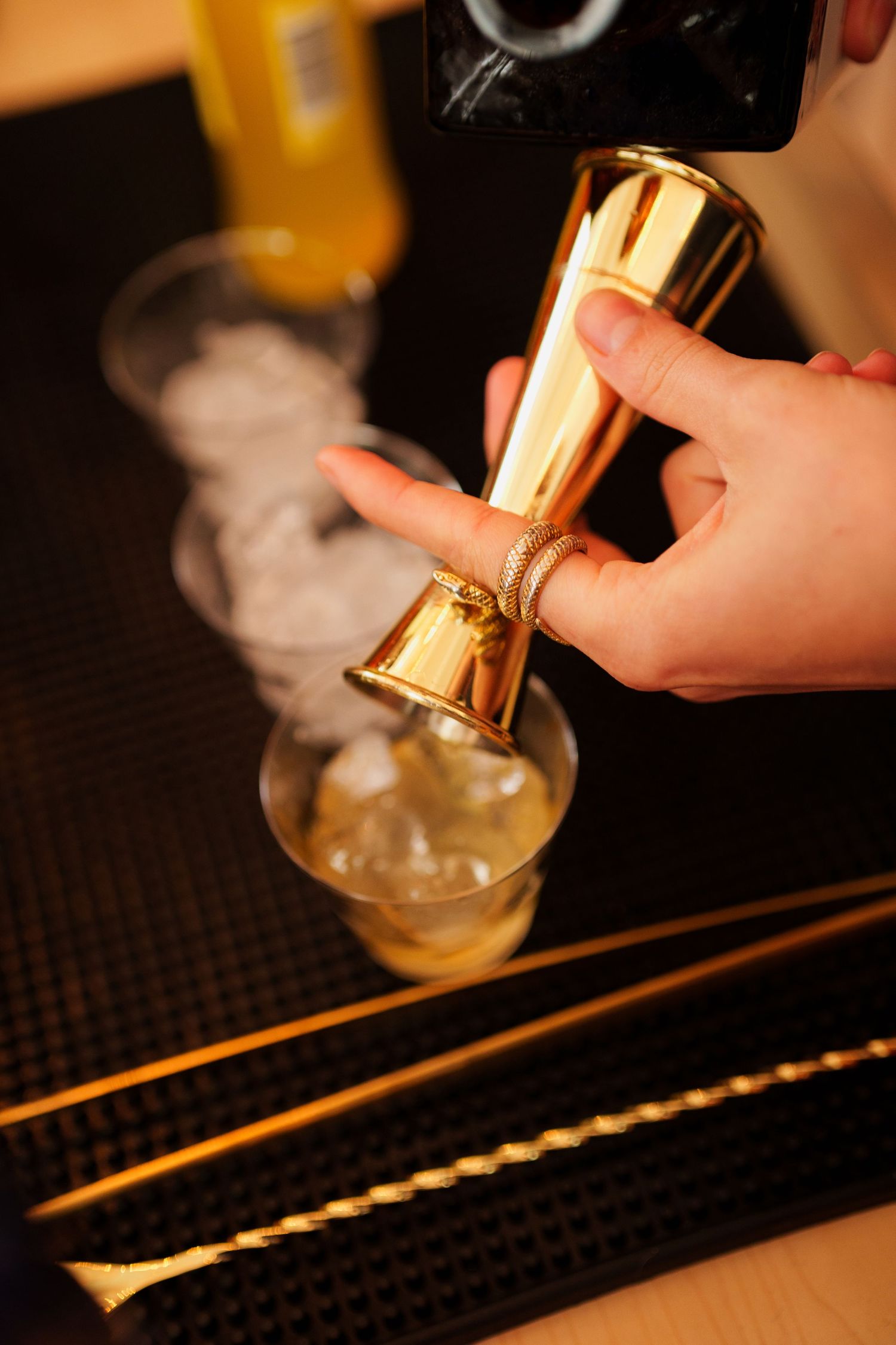Close-up of hands using a gold jigger to measure cocktail ingredients on a bar mat.