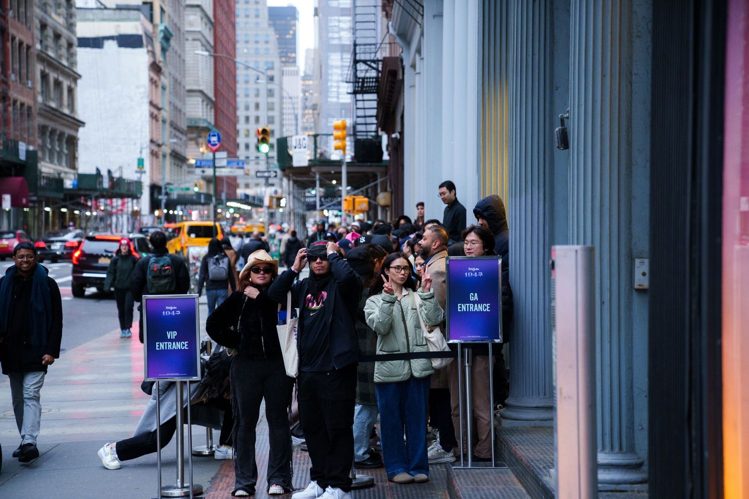 Queue of shoppers along a city sidewalk with stanchions and yellow taxis in background.