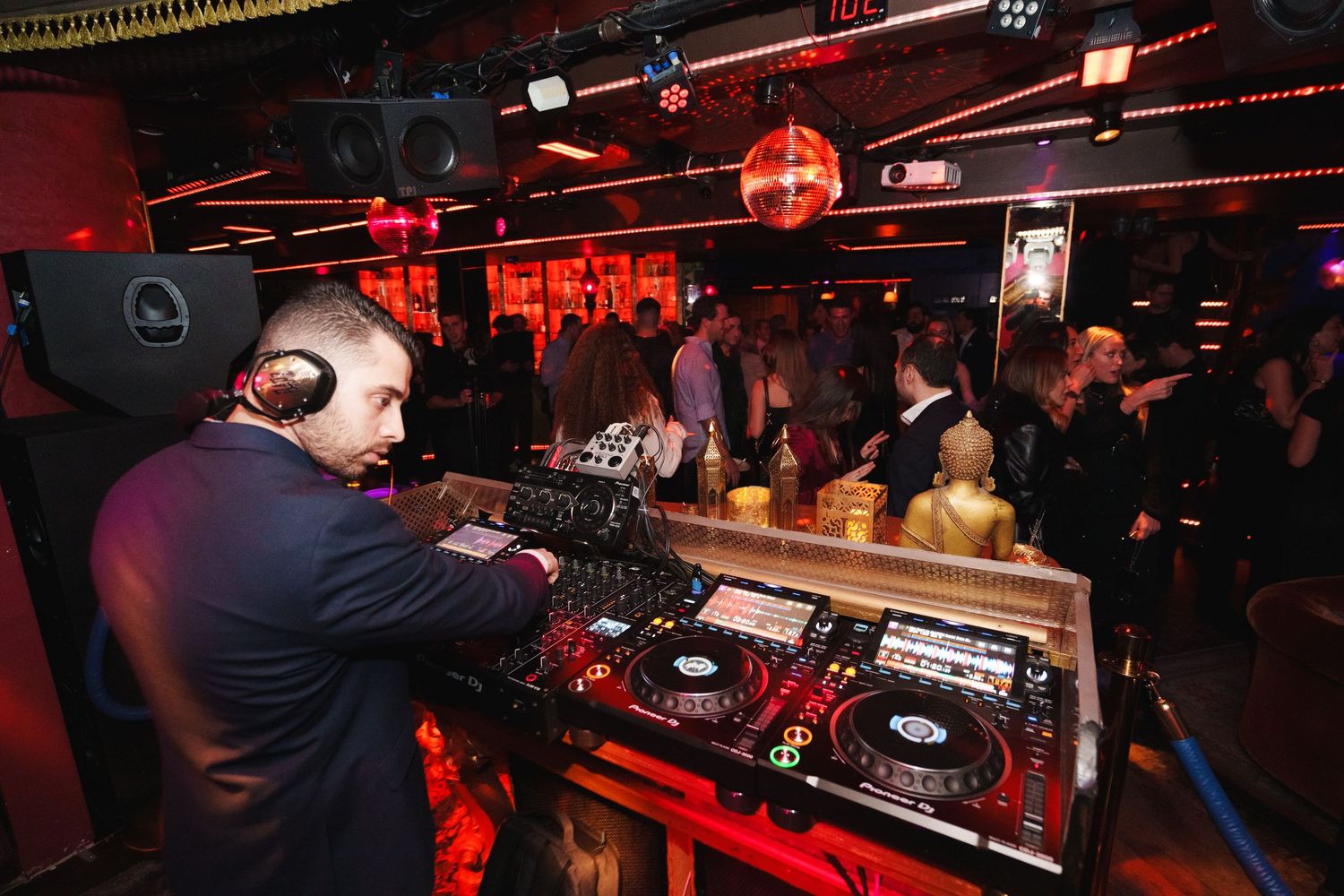 DJ working the turntables at a crowded nightclub with red ambient lighting.