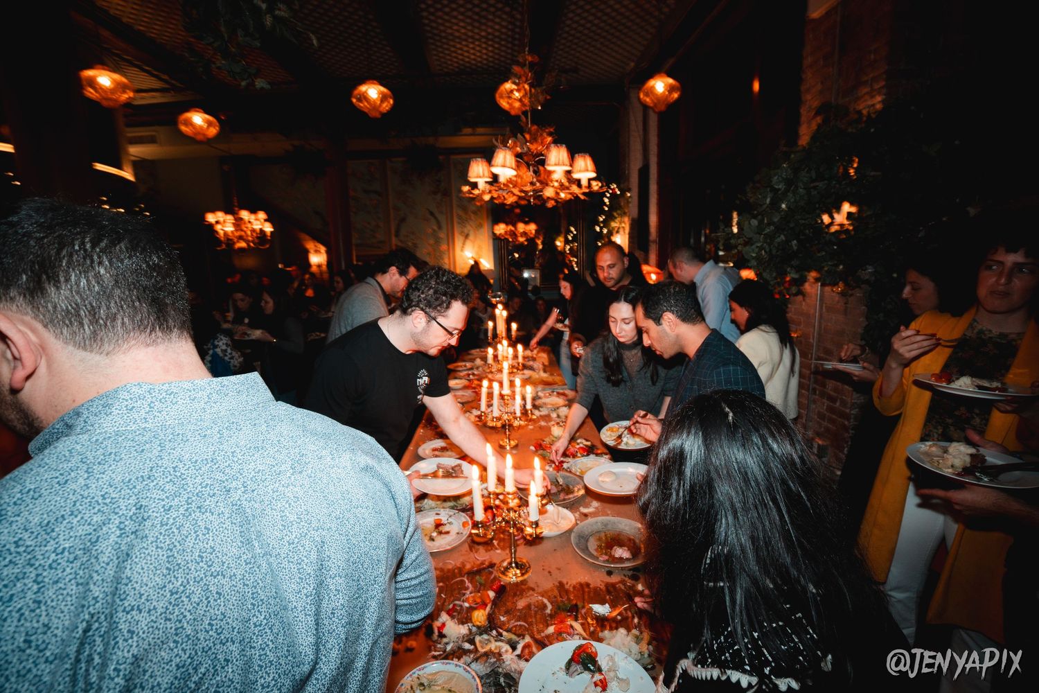 Guests gather around a candlelit table at a dimly lit dinner party with warm ambient lighting.
