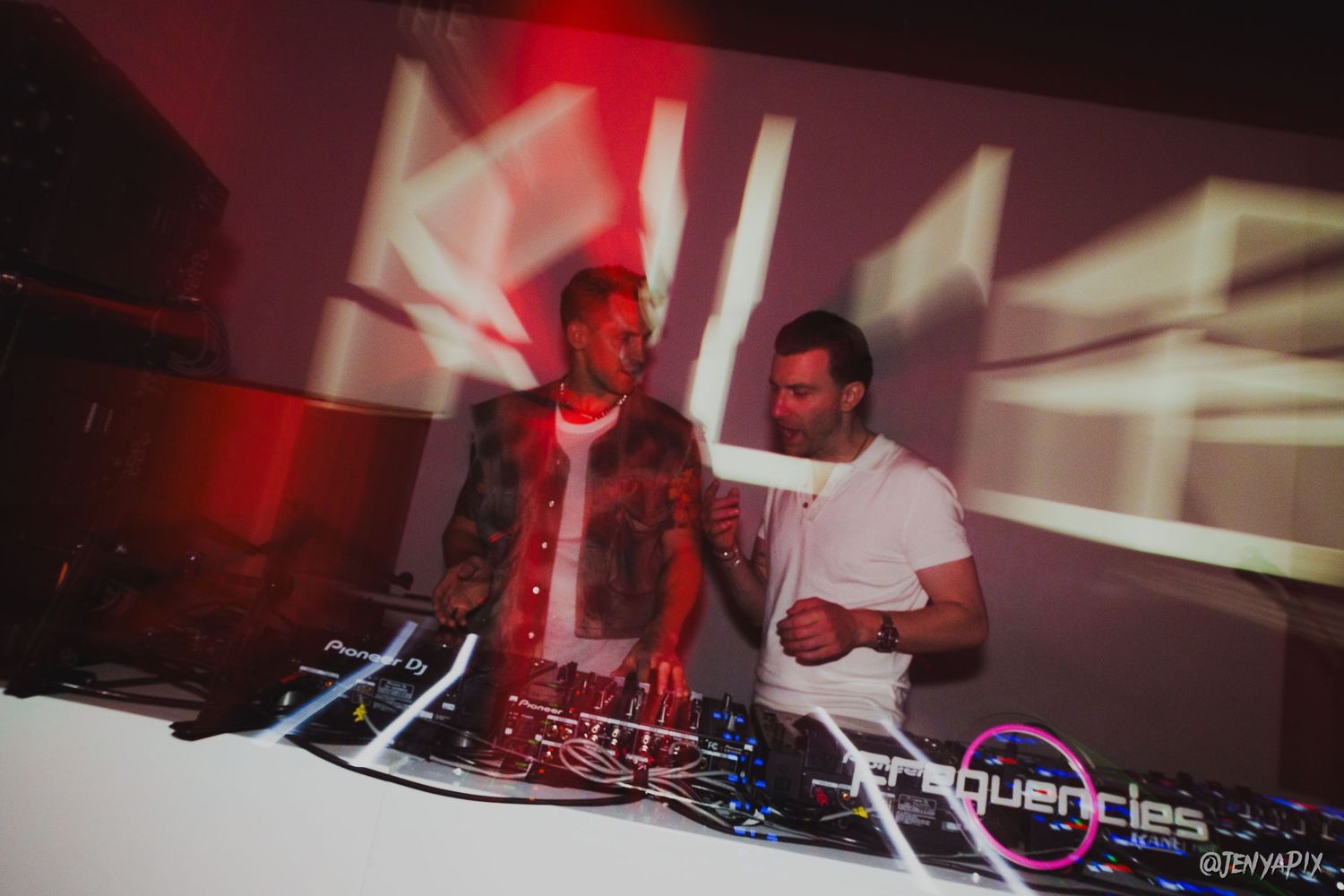 Two DJs mixing music at a club booth with red lighting and projected visuals.