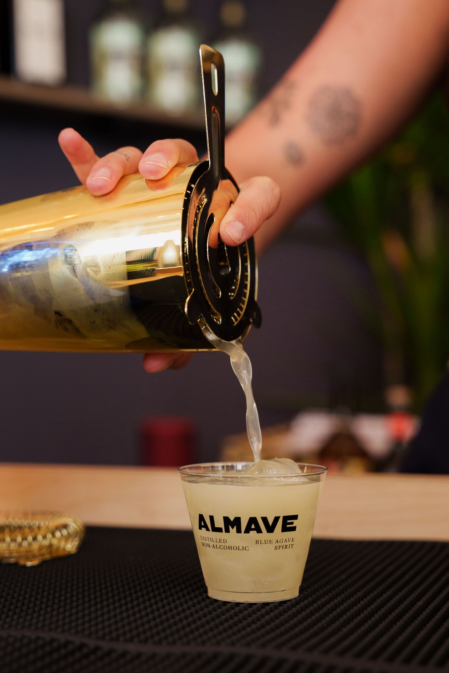 Clear liquid being poured from a gold shaker into an Almave branded plastic cup.