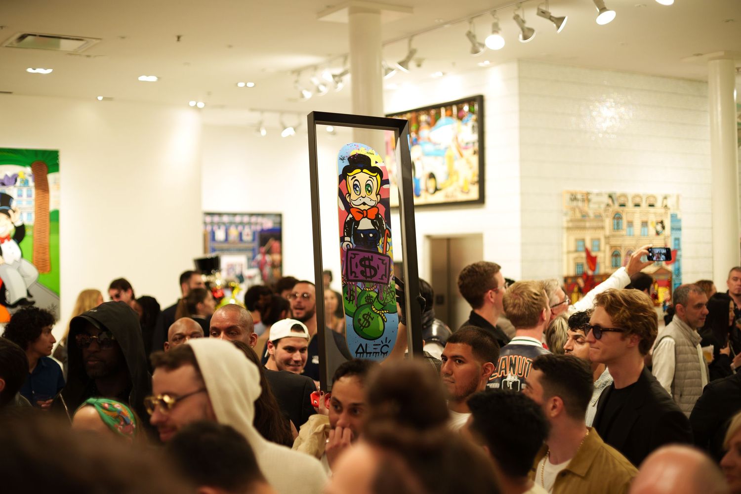 Crowded art gallery opening with colorful framed artwork on white walls and track lighting overhead.