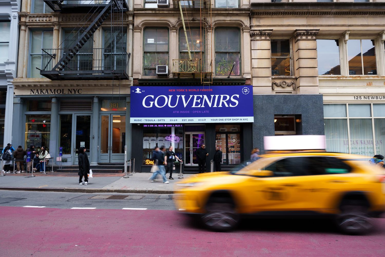 Yellow taxi drives past a neon 'Gouvenirs' storefront sign on a busy city street.