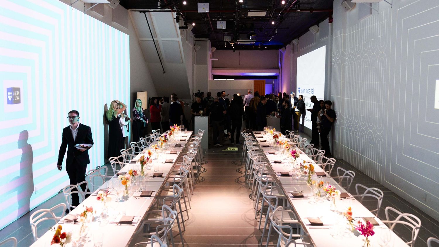 Long white dining tables setup in minimalist event space with projection lighting.