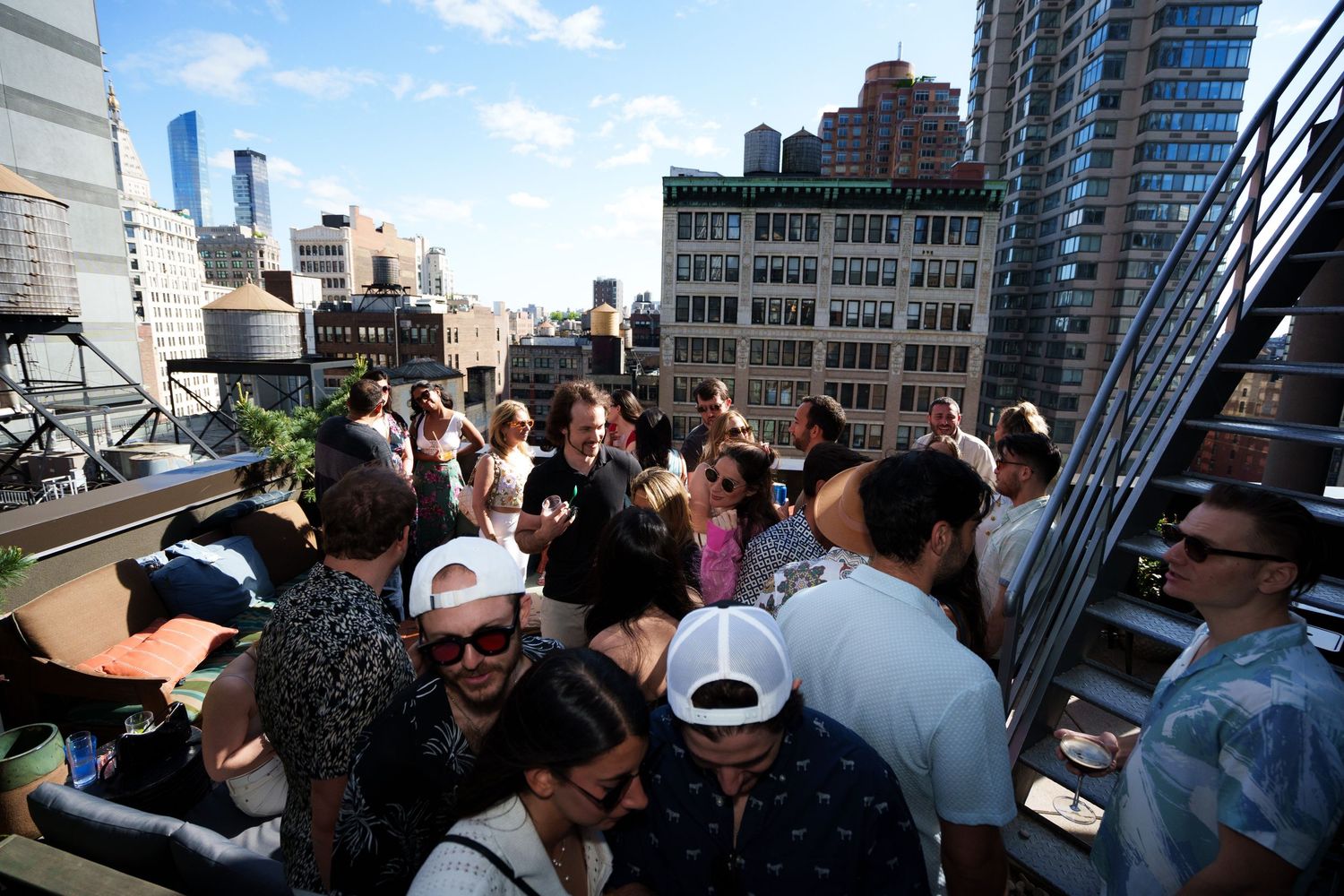 People socializing at crowded rooftop event with city buildings in background.