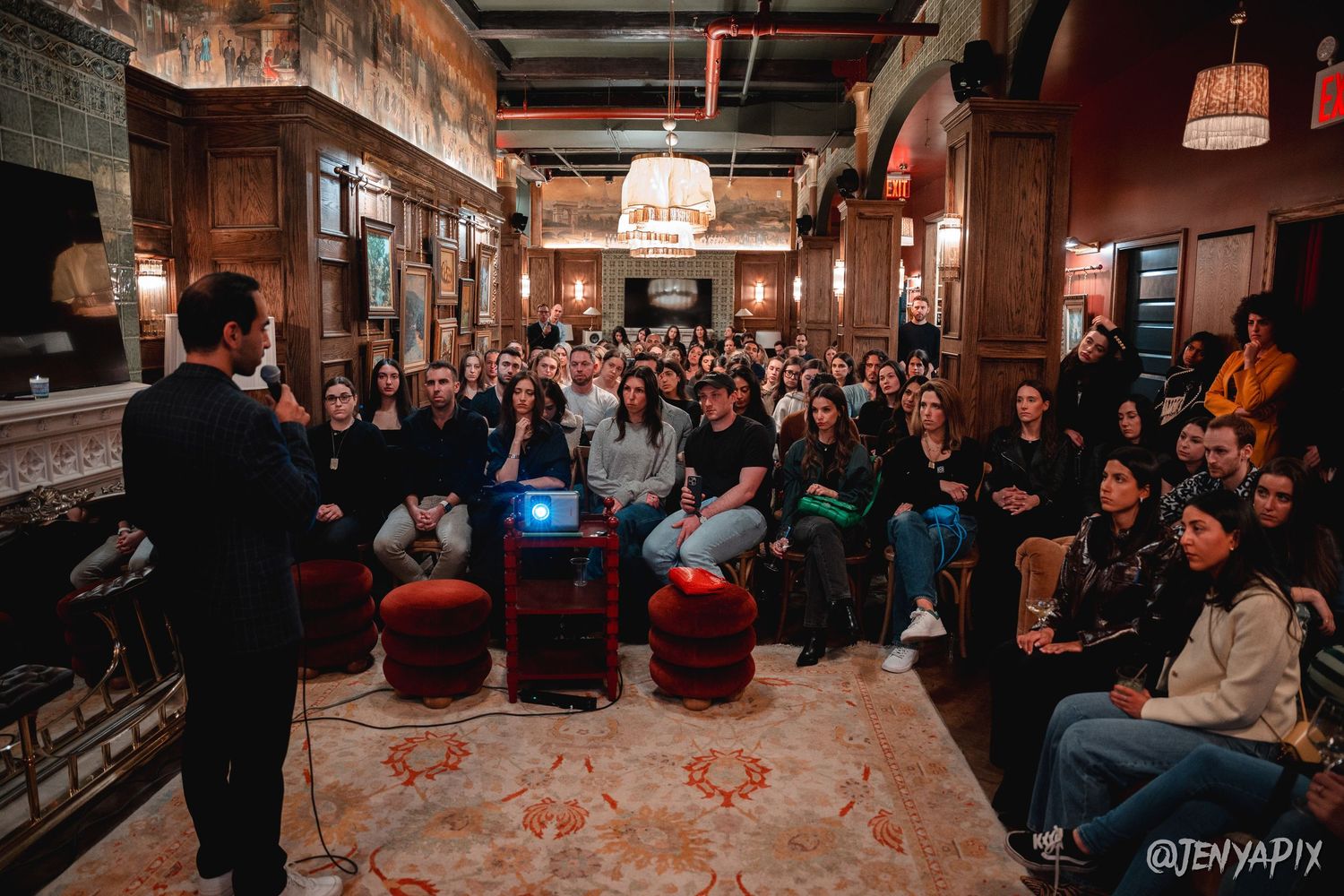 Indoor event venue with audience seated facing presenter in wood-paneled room.