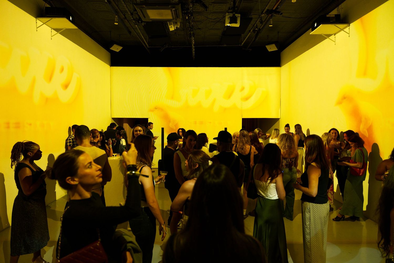 Yellow-lit gallery walls extend down a hallway filled with art viewers attending an exhibition opening.