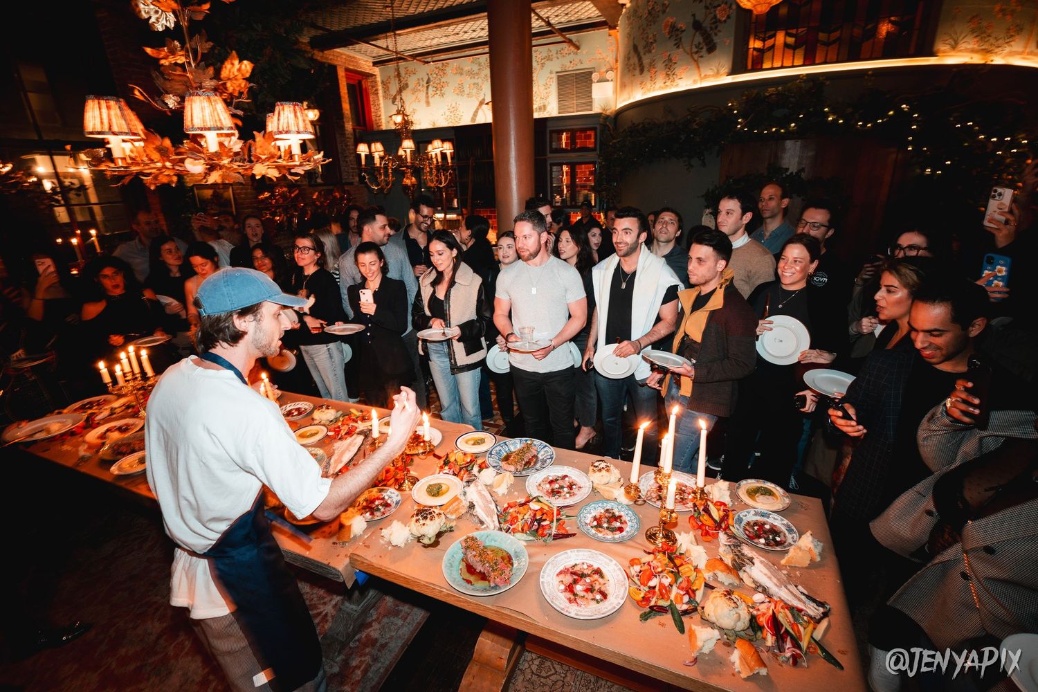 Crowd gathered around a food presentation table at an evening event.