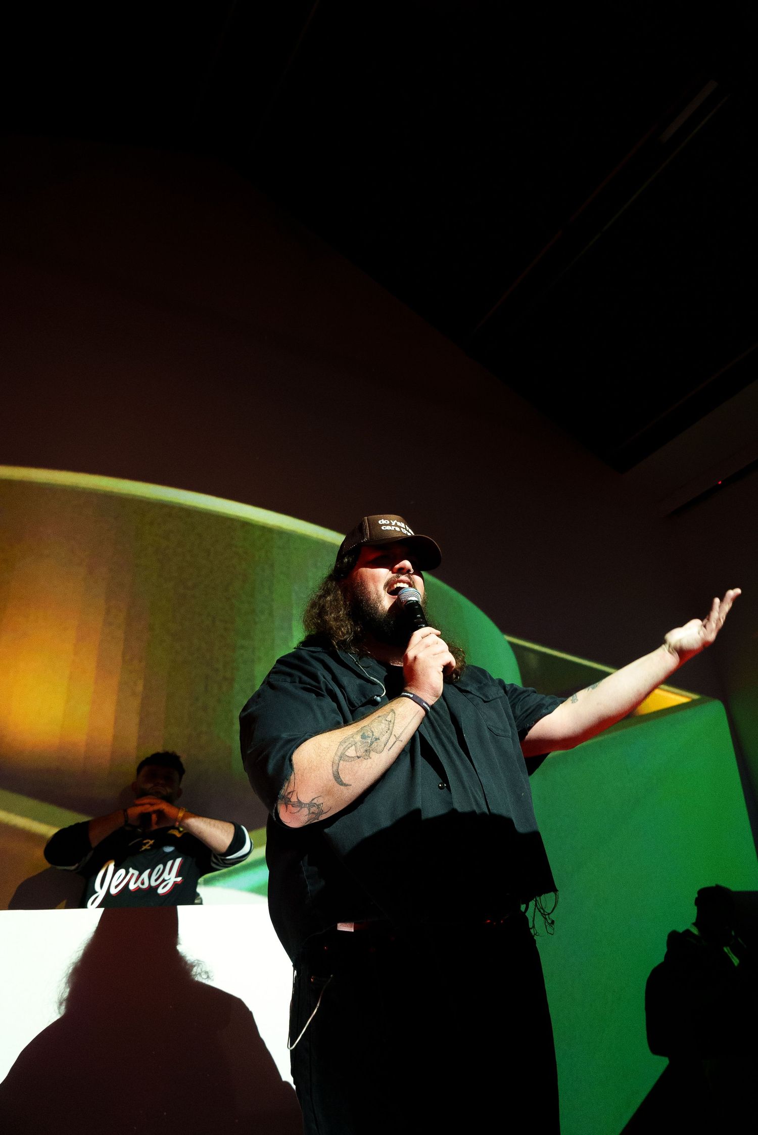 Performer in dark clothing gestures while singing on stage with green lighting effects.