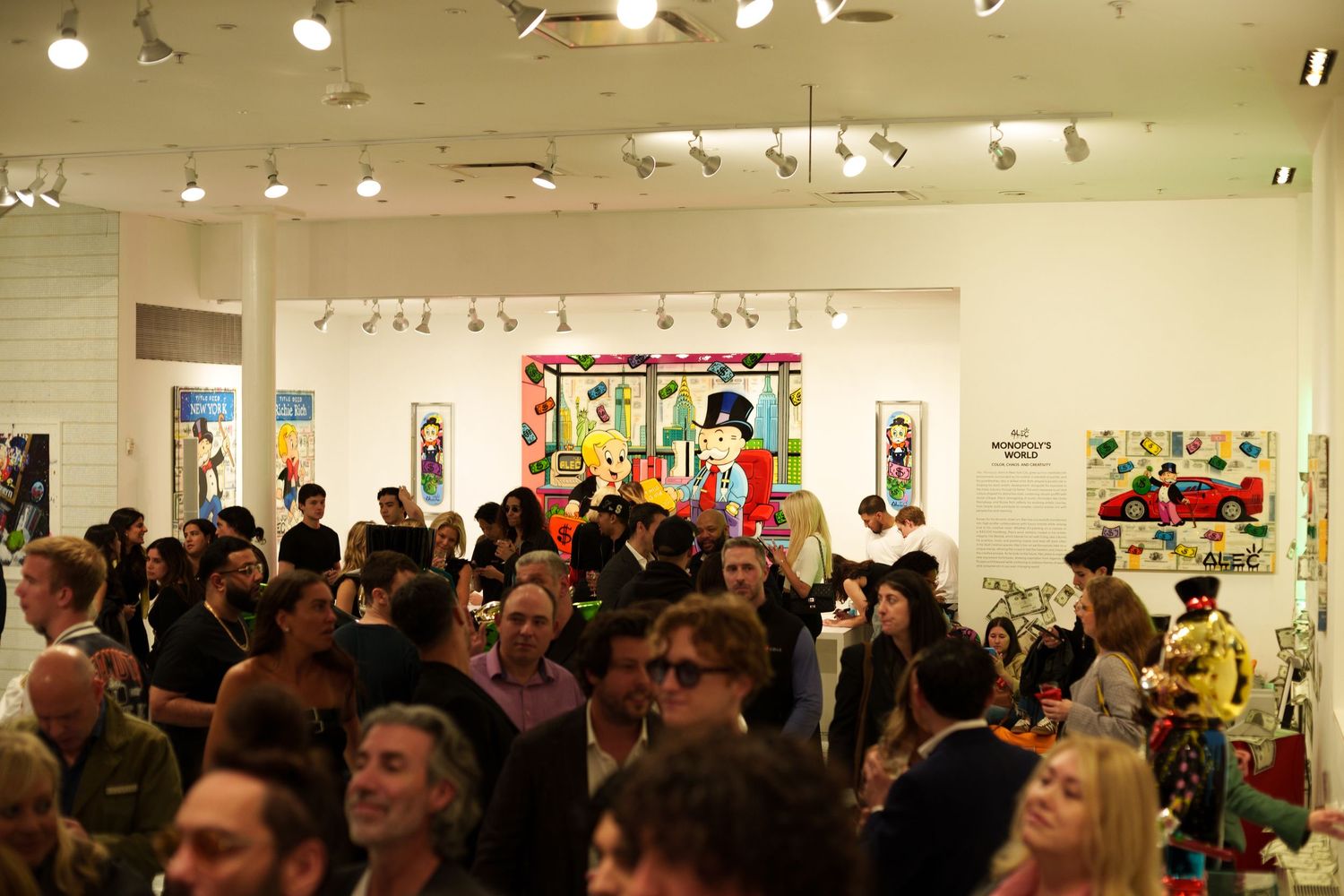 Packed art gallery opening with bright overhead lighting and colorful artwork on walls.