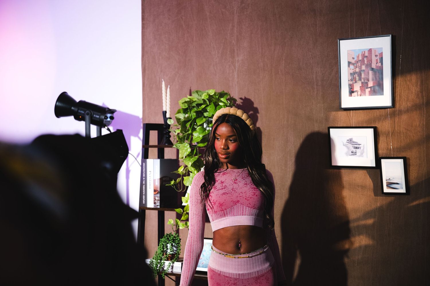 A model in a pink crop top poses during a professional photo shoot with lighting equipment and greenery.