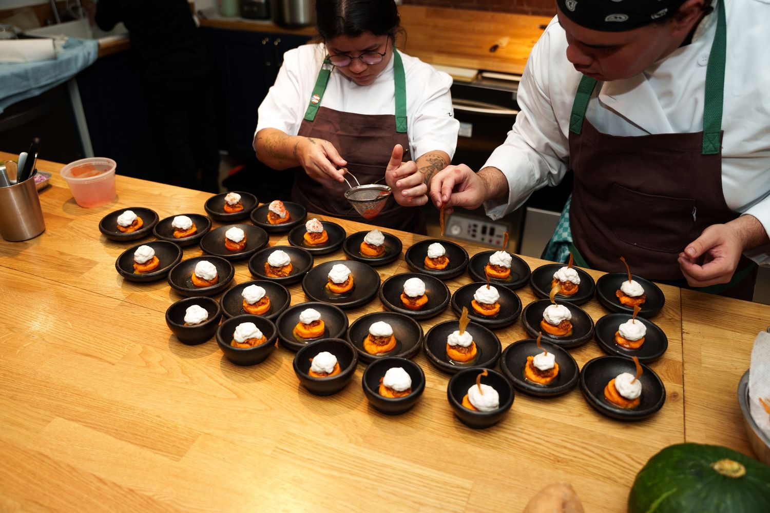 Chefs plating desserts on multiple dark plates arranged on a wooden table in a professional kitchen setting.