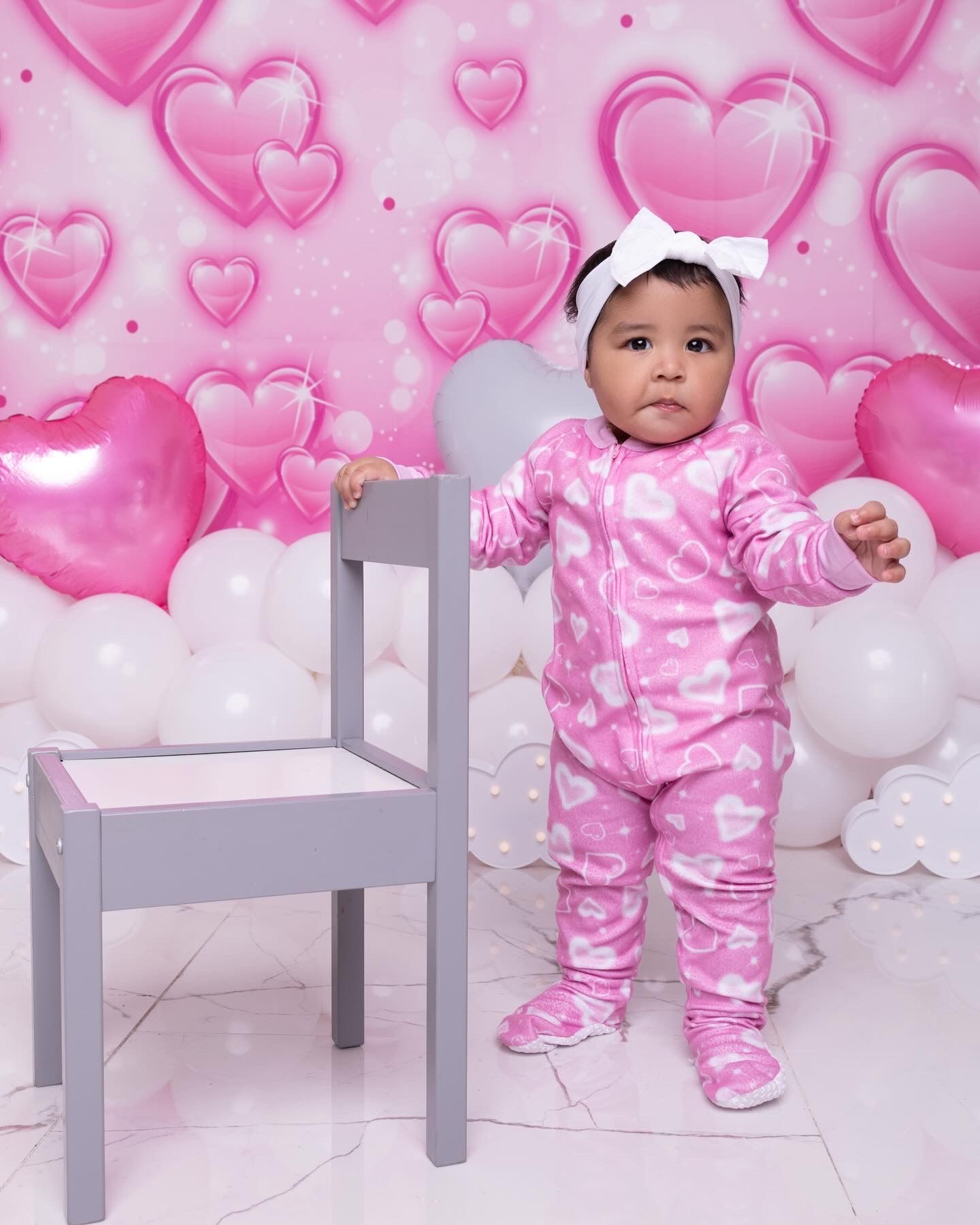 A baby in pink heart patterned pajamas stands next to a gray chair against a pink heart backdrop.