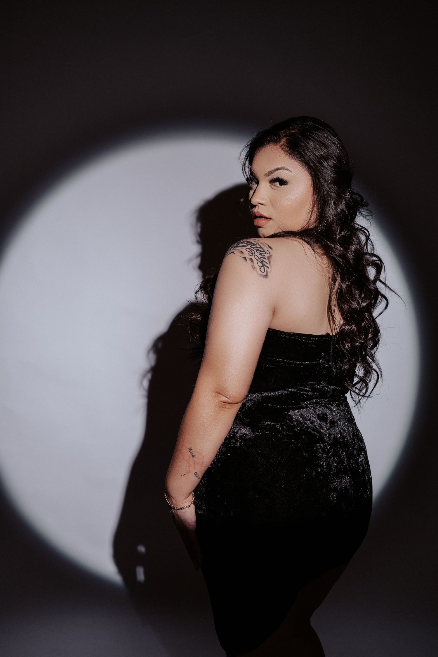 Dramatic portrait in black velvet dress with spotlight effect and visible tattoo details.