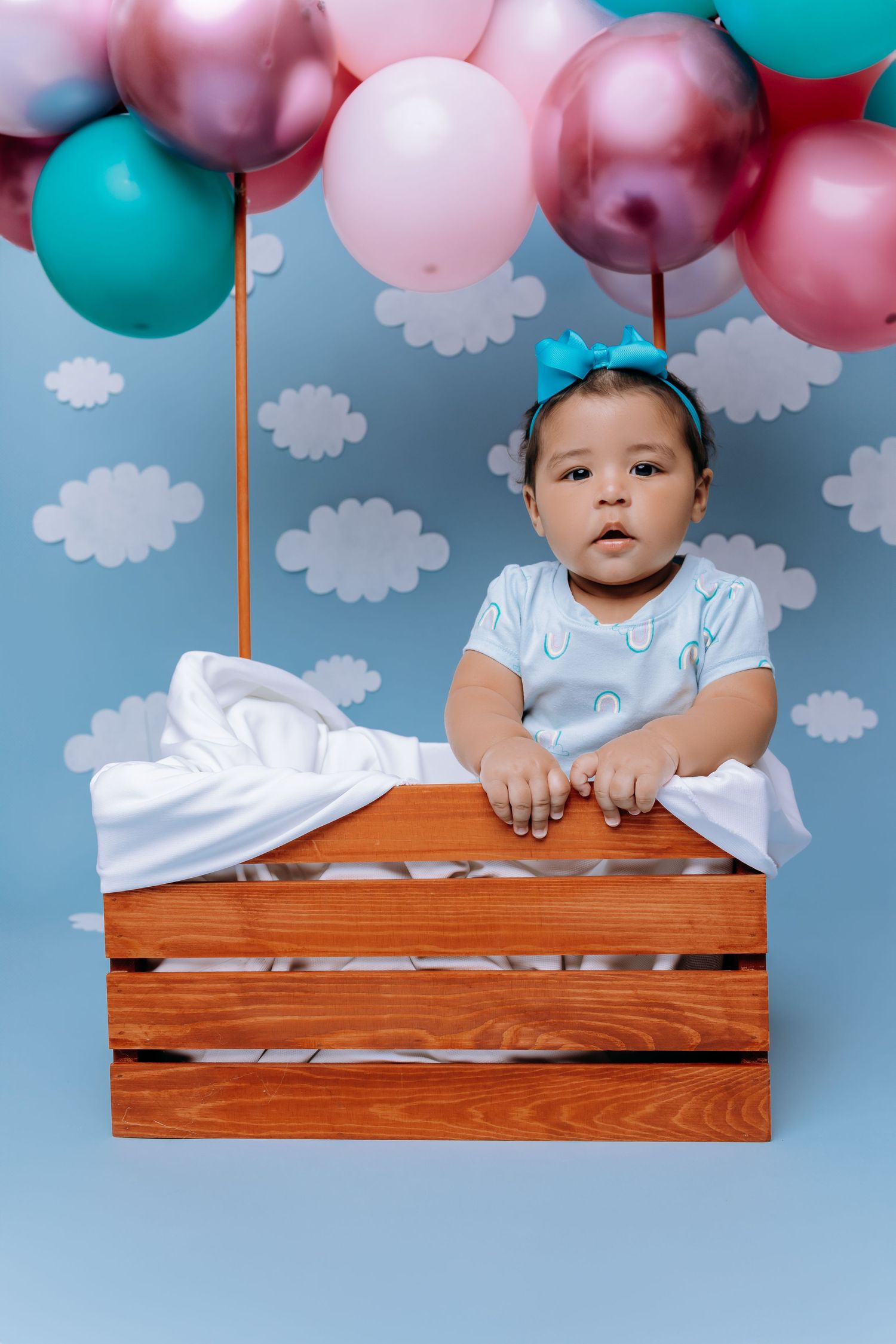 A baby sits in a wooden crate with pink and teal balloons against a cloudy backdrop.