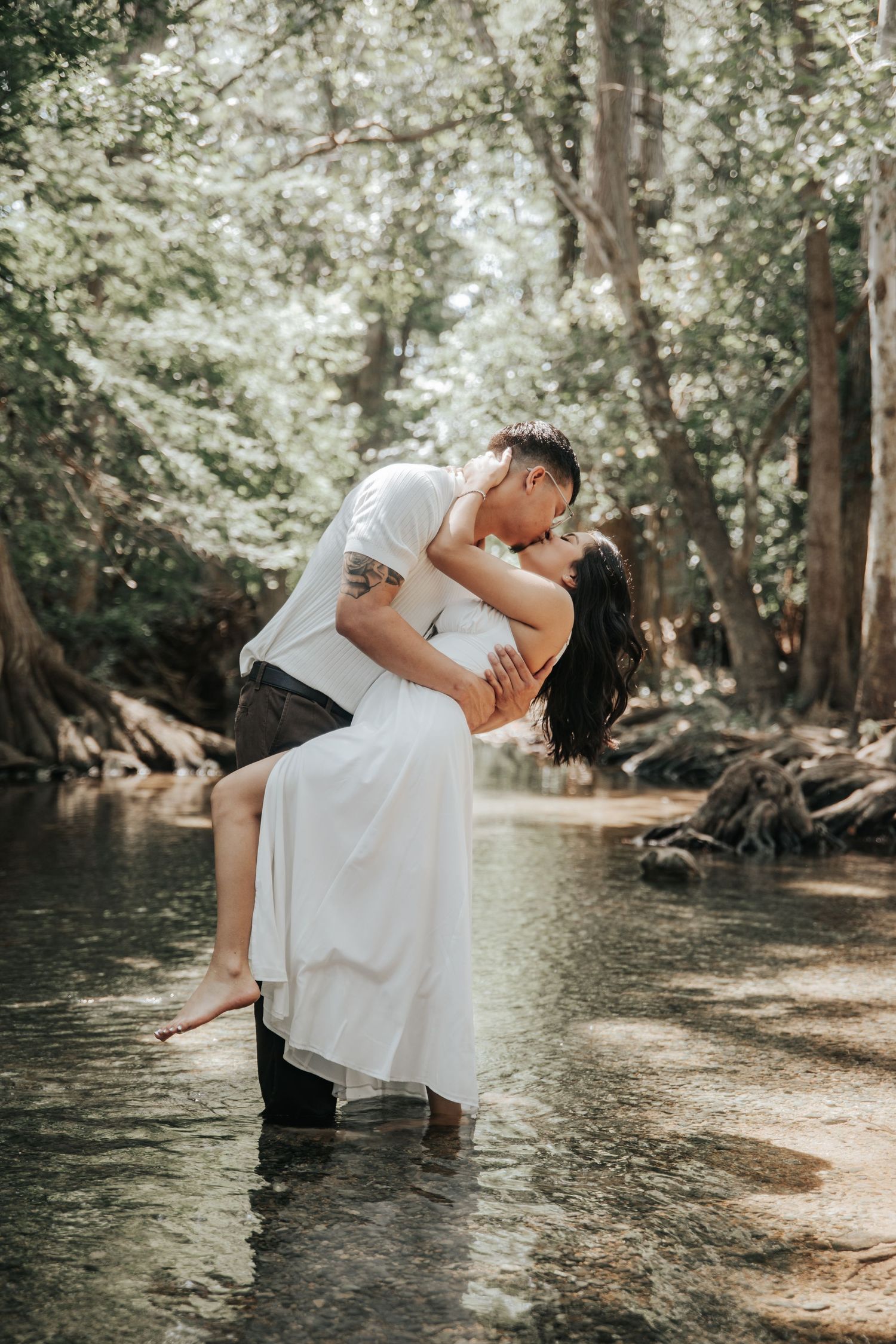 A romantic couple embraces in a dip kiss while standing in shallow creek waters surrounded by sunlit forest trees.