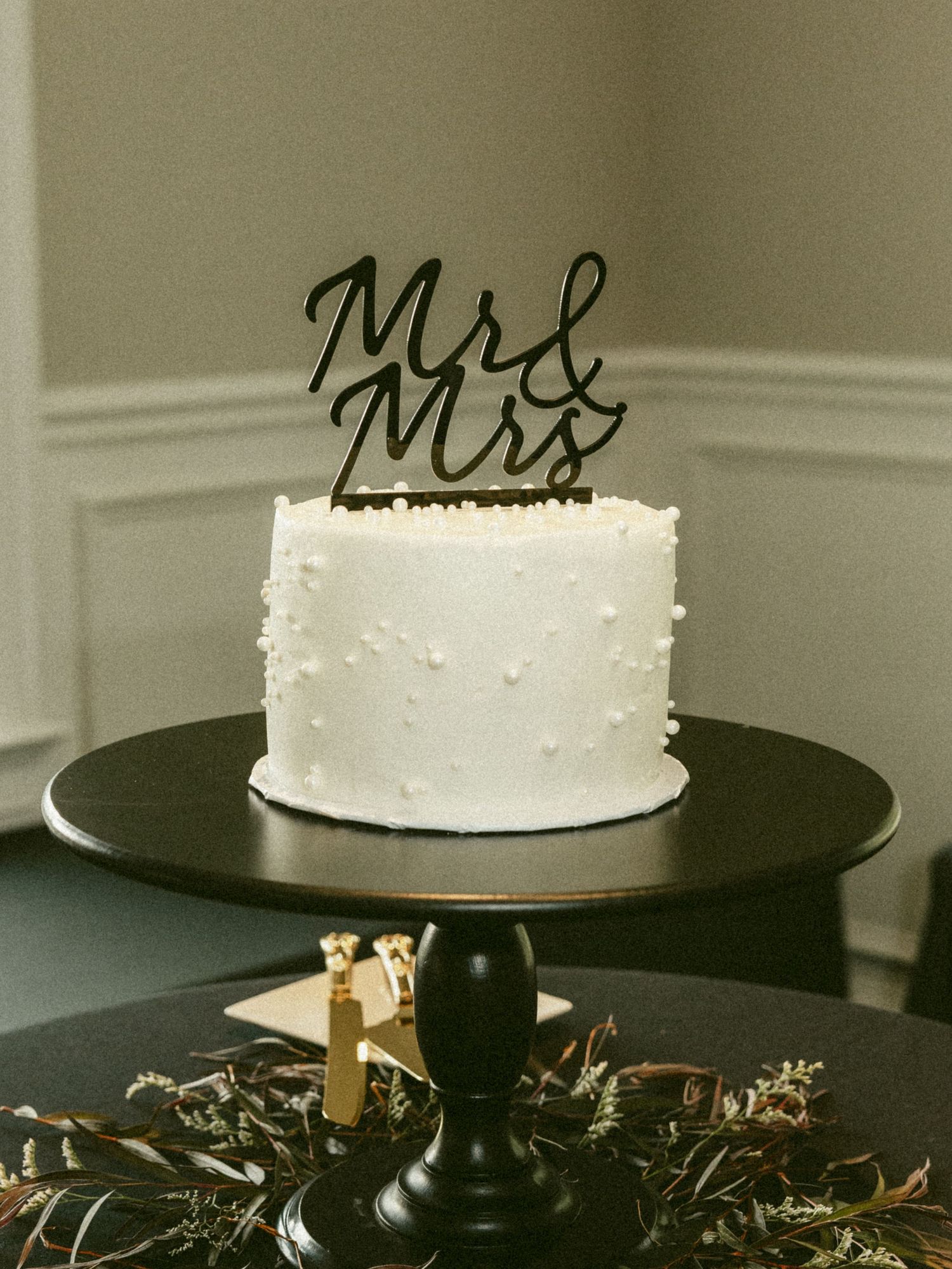 A minimalist white wedding cake displayed on a black pedestal stand with delicate greenery accents. Located in Bellbrook, Ohio.
