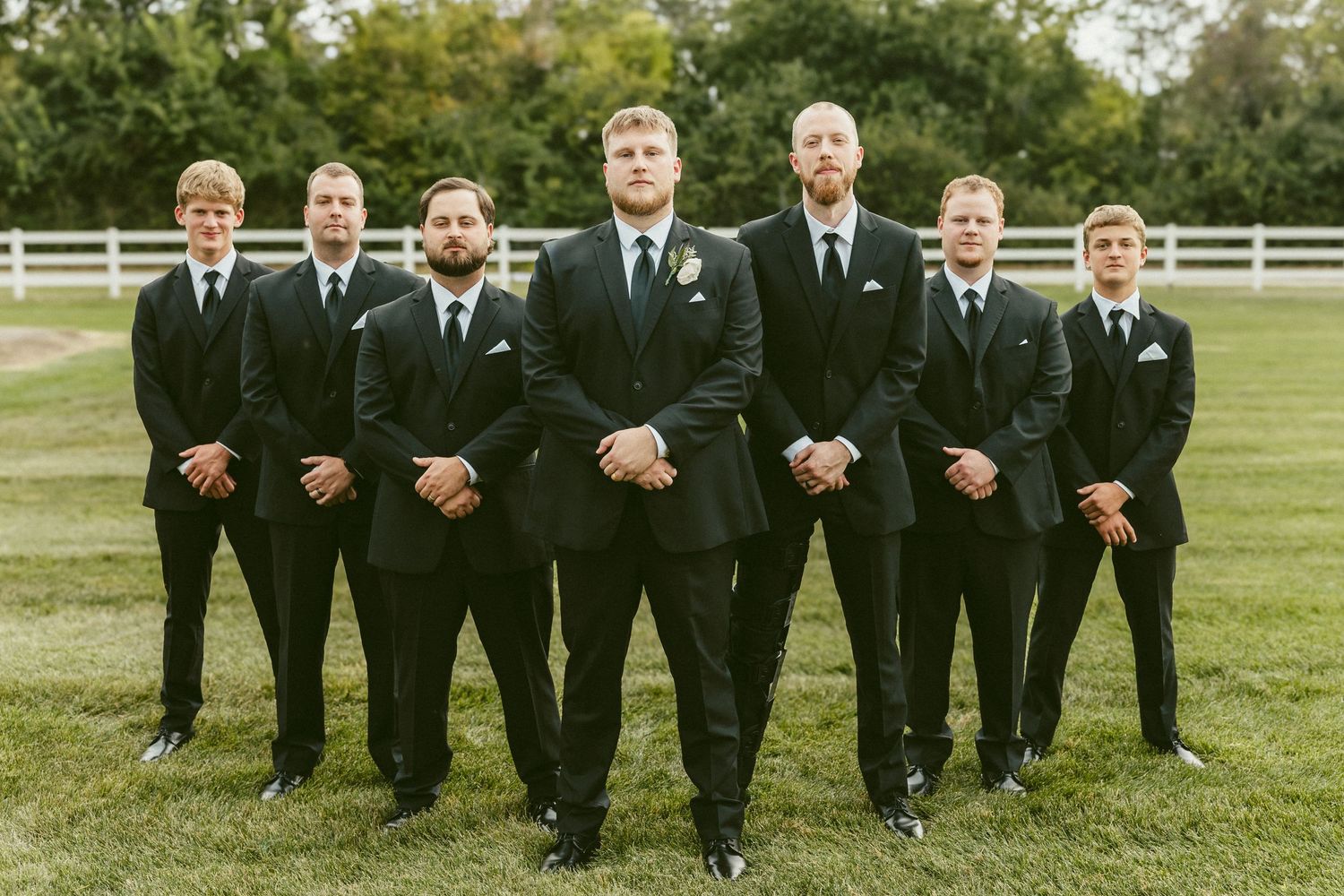 Groomsmen in black suits walk in a line formation across a lush green lawn on a sunny wedding day. Located in Bellbrook, Ohio.