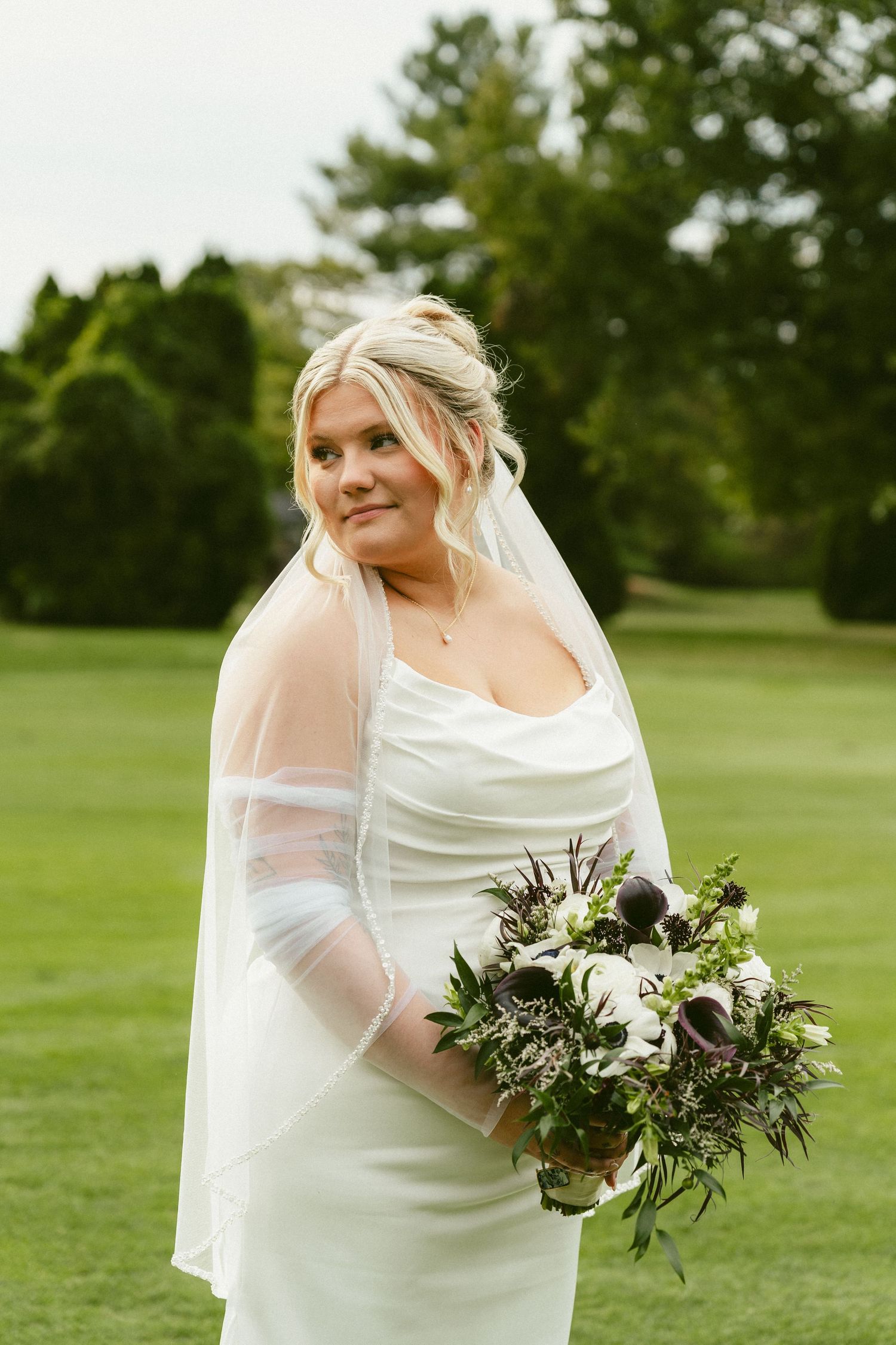 A bride in a white fitted dress with long veil holds a lush green and white bouquet on a golf course. Located in Bellbrook, Ohio.