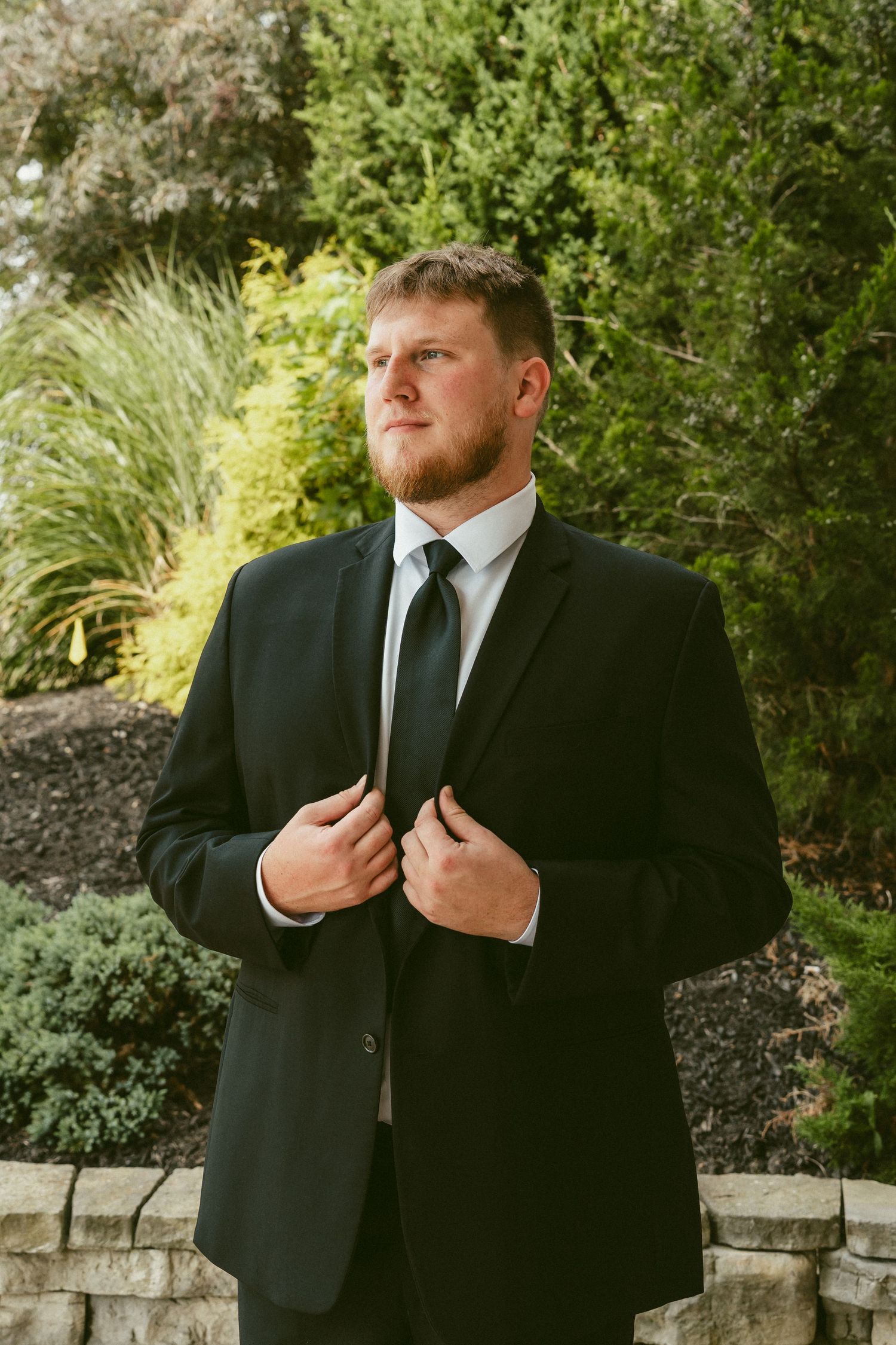 A groom in a black suit adjusts their tie while standing in a garden setting with lush greenery in the background Located in Bellbrook, Ohio