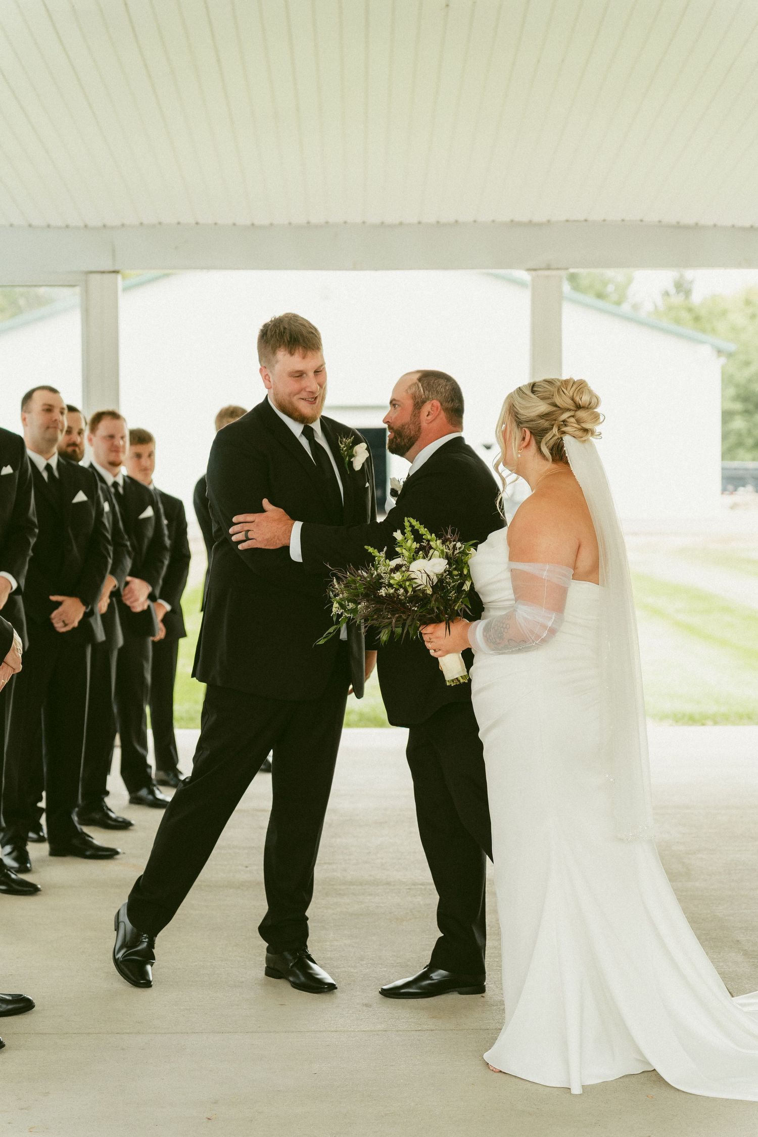 Wedding party members in black suits and white dress gather for formal photos under a covered outdoor venue. Located in Bellbrook, Ohio.