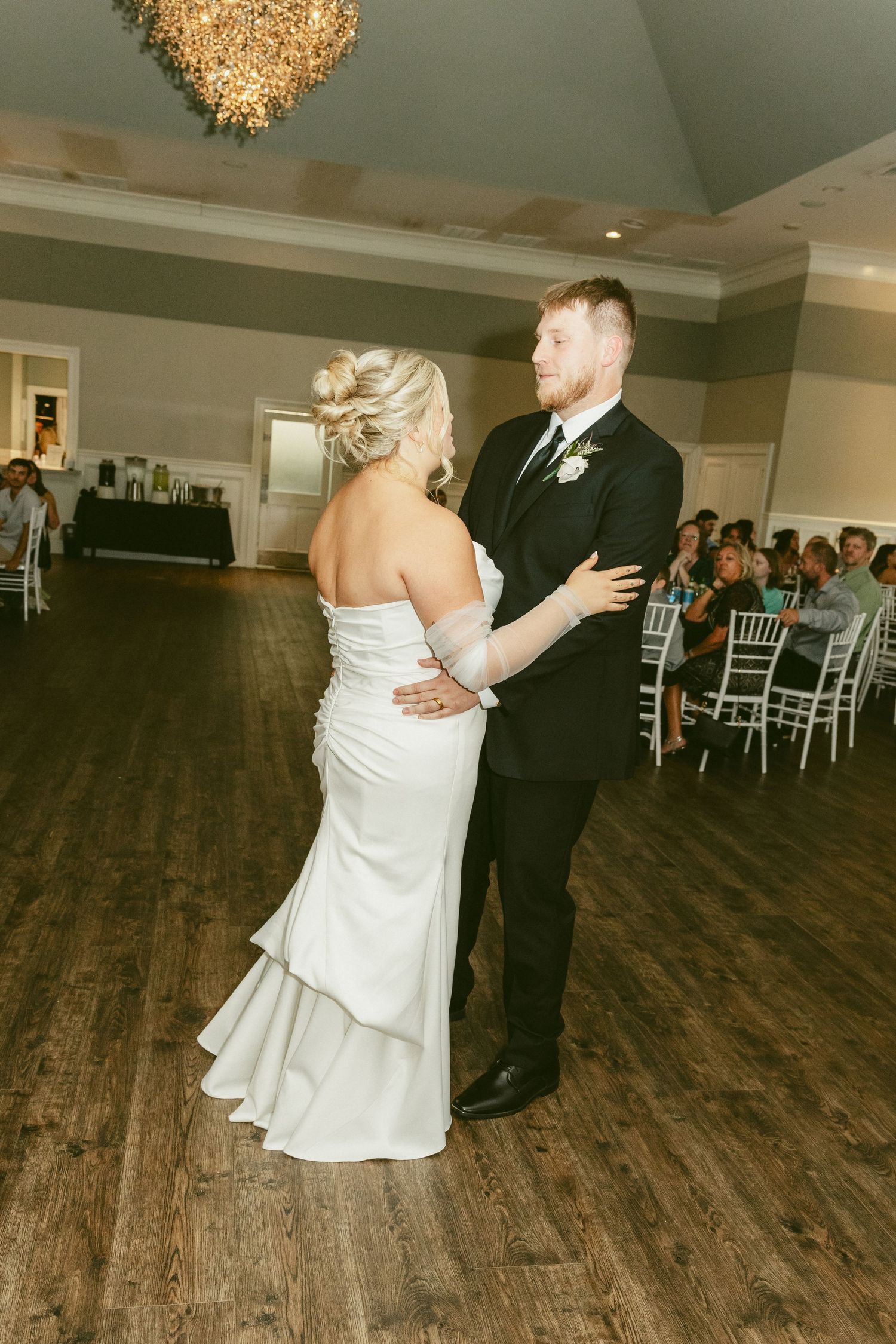 A couple shares their first dance as newlyweds on a wooden dance floor beneath an elegant crystal chandelier. Located in Bellbrook, Ohio.